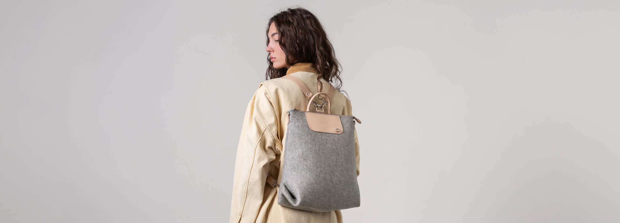 Person in a long-sleeved, cream-colored dress holding a gray, perforated handbag with a wooden handle, standing against a textured rock background.