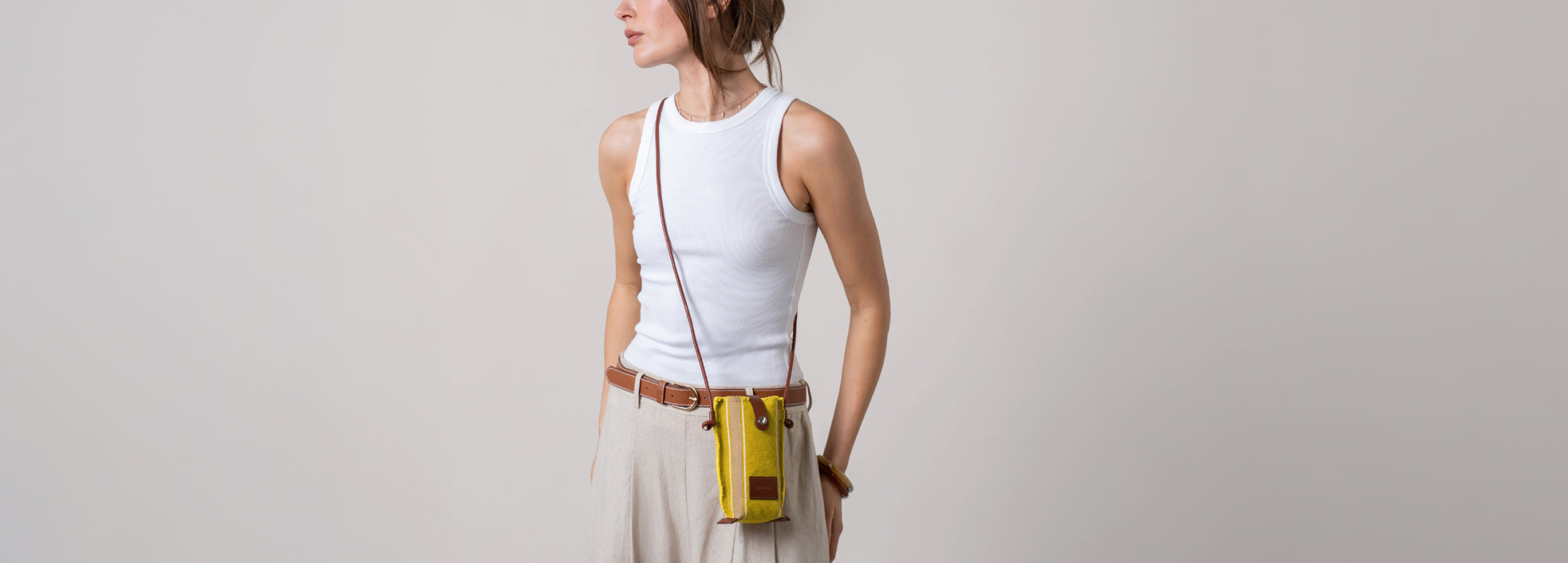 A person in a light beige outfit holding a brown crossbody bag with a beige flap, adjusting the strap. The surroundings are a neutral, soft background.