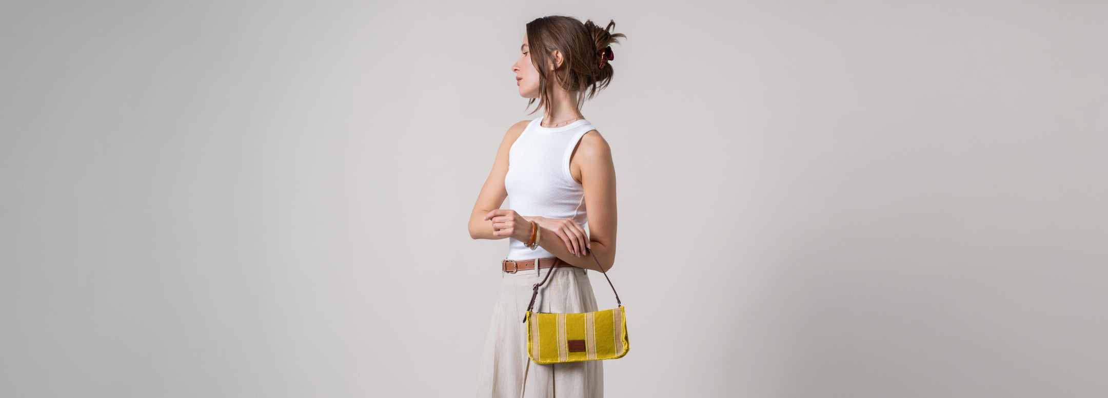A woman stands in front of a circular wall feature, looking at her phone. She wears a plaid jacket, white shirt, brown skirt, and holds a red and black handbag.