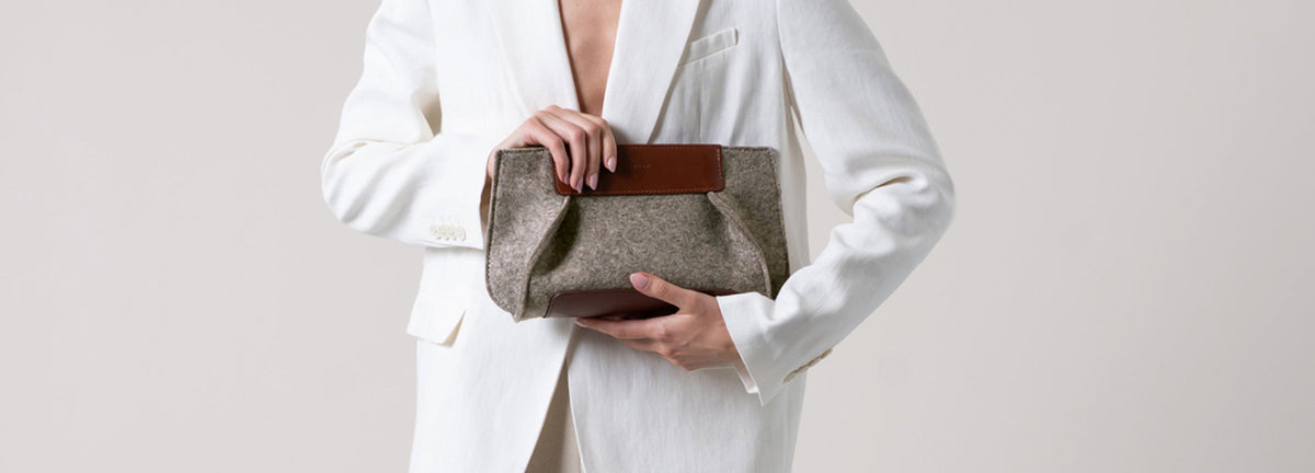 Person in a white blazer holds a gray and brown clutch bag against a plain light background. The face is not visible, focusing on the outfit and accessory.