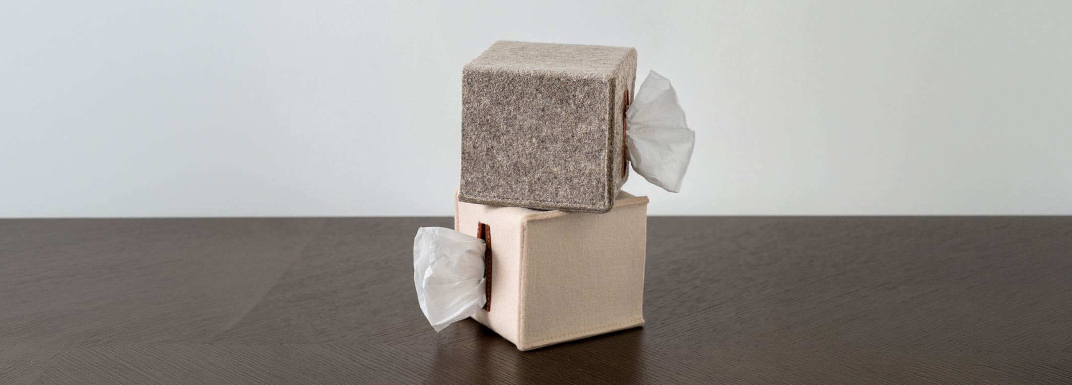 A grey felt tissue box sits on top of a book titled MARK BRADFORD, placed on a wooden table with blurred objects in the background.