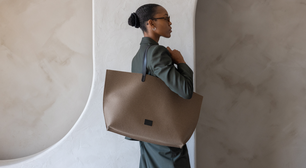A woman in a green suit carries a large brown tote bag over her shoulder, standing in front of a textured, light-colored wall with decorative curves.