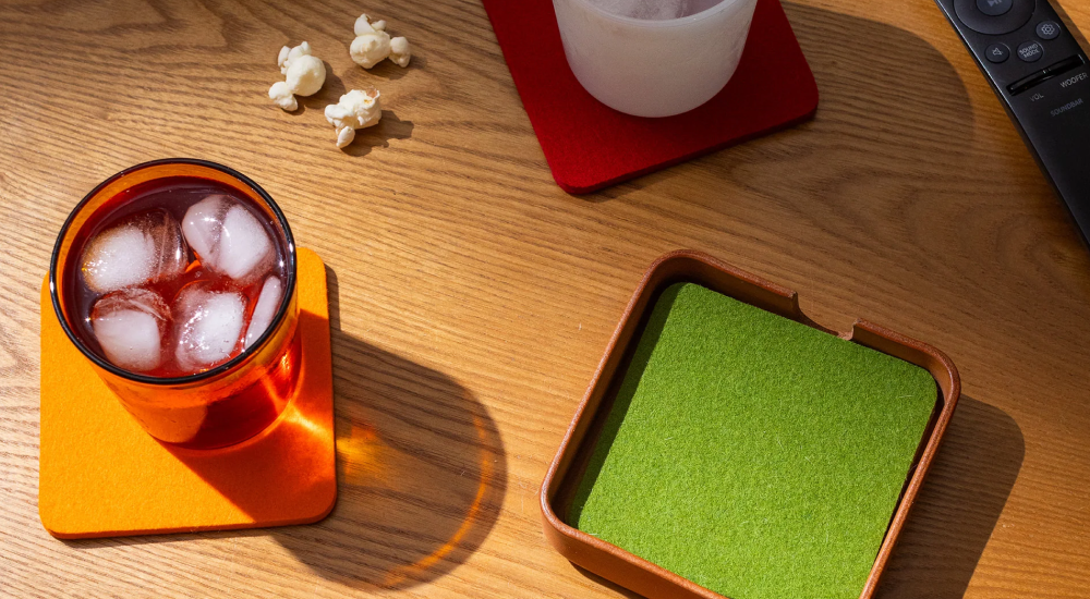 A glass of iced red drink on an orange coaster, a white cup on a red coaster, a green coaster in a holder, a few popcorn pieces, and a remote control on a wooden table in sunlight.