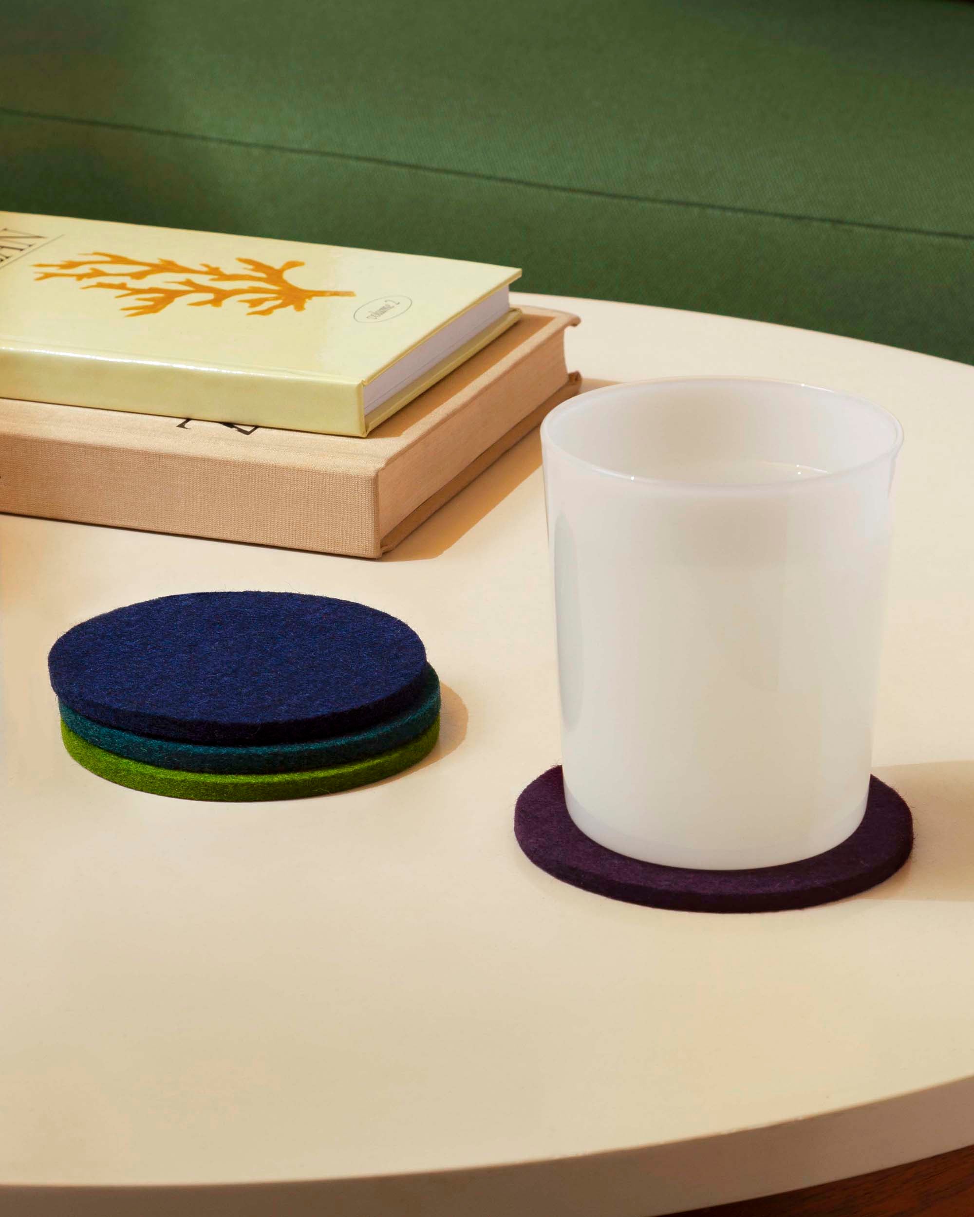 A white glass sits on a purple felt coaster on a round table, next to a stack of colorful felt coasters and a green book with a coral design on top of a closed box.