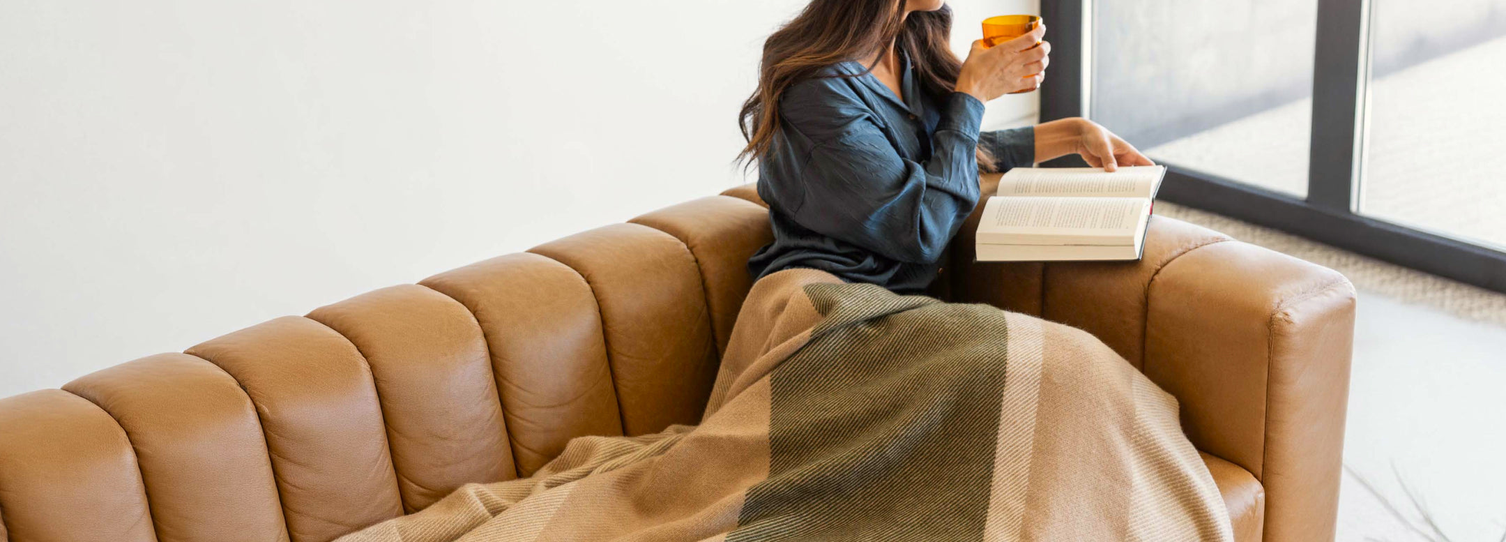 A woman sits on a tan couch by a window, wrapped in a blanket, holding an orange mug and an open book, appearing relaxed and comfortable.