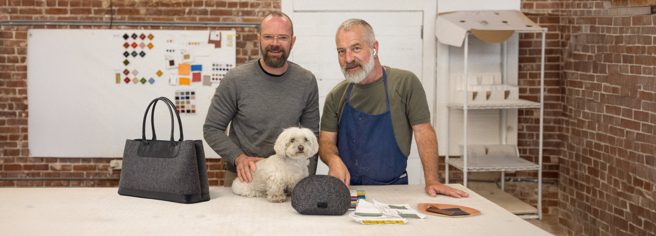 Two smiling men stand at a worktable with a small white dog, bags, and fabric samples in a studio with brick walls and shelves in the background.