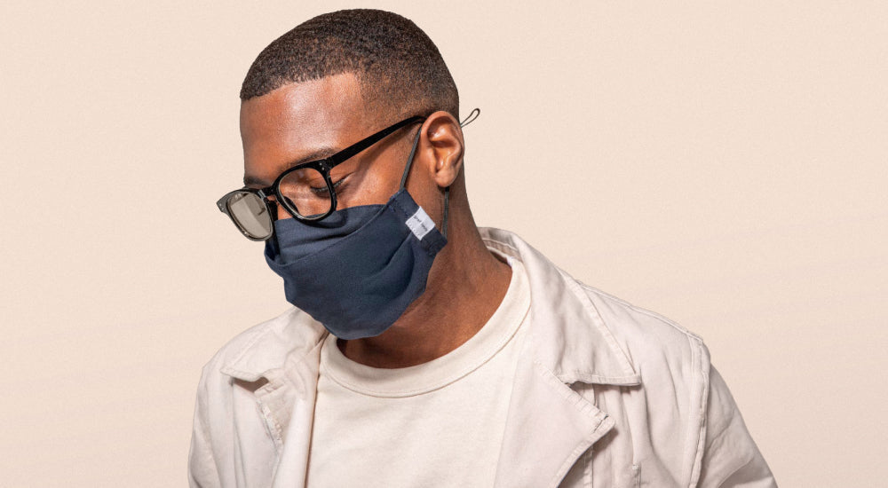 A man wearing glasses, a navy blue face mask, a white t-shirt, and a light jacket looks downward against a plain beige background.