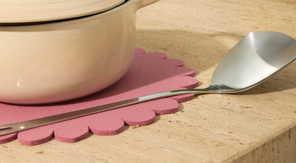 A cream-colored pot sits on a pink scalloped felt trivet next to a metal spatula on a beige countertop.