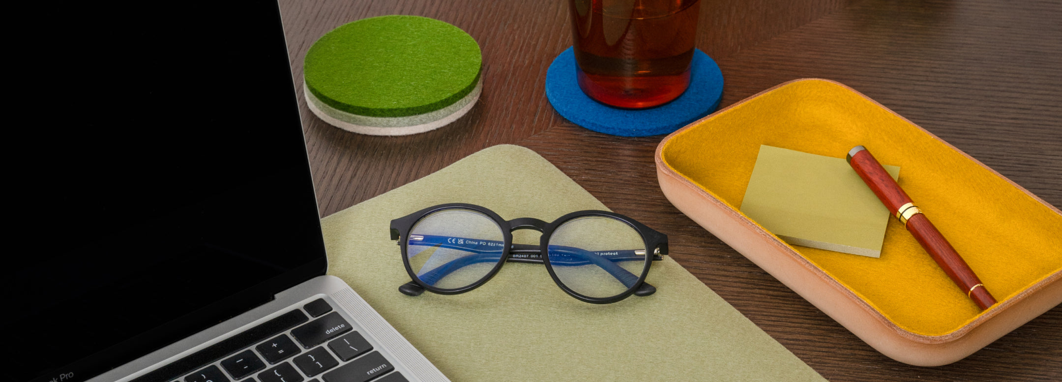 A hand using a white mouse on a blue desk mat next to a keyboard, a mug of coffee on a red coaster, and a yellow tray holding office supplies.
