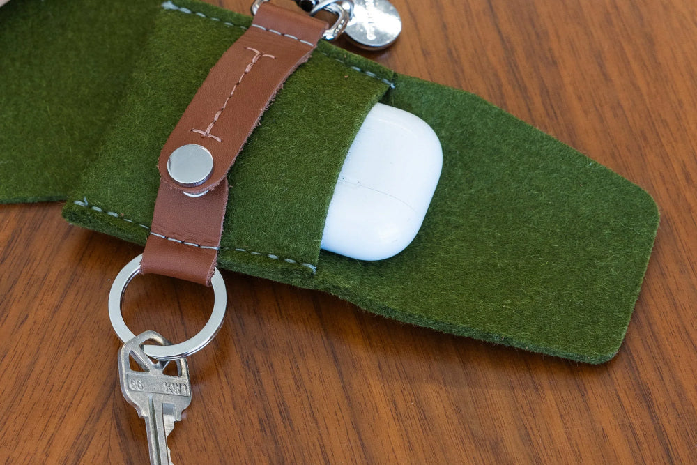 A green felt keychain pouch with a brown leather strap holds a set of keys and a white wireless earbud case, resting on a wooden surface.