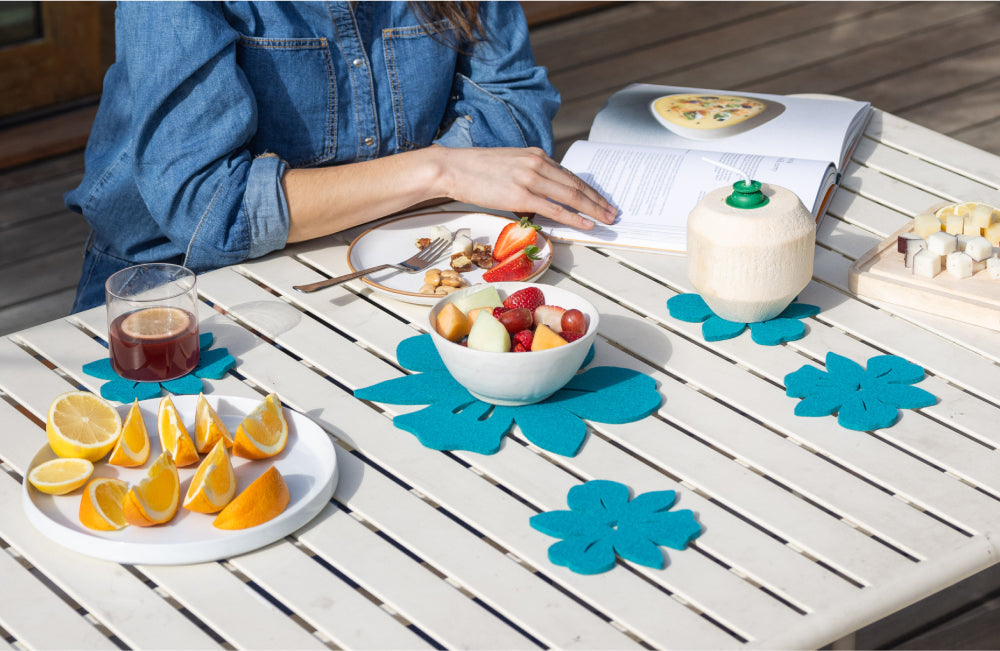 A person in a denim shirt sits at a table with fruit, a drink, a coconut, snacks, and an open cookbook. Blue flower-shaped mats decorate the white table.