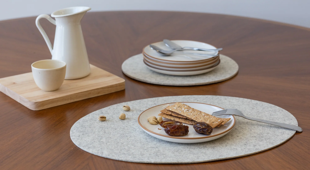 A round wooden table set with a plate of crackers, dates, and nuts, a fork, stacked plates with cutlery, and a white pitcher and cup on a wooden board.