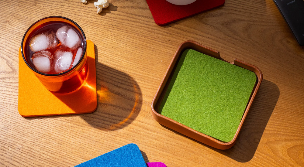 A beige ceramic mug on a red coaster sits on a white table, alongside red coasters, an orange notebook, a wooden clock, and a green potted plant.
