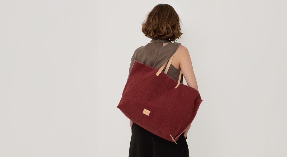 A woman with short brown hair, wearing a sleeveless dark top and skirt, is seen from behind carrying a large maroon tote bag with tan handles over her shoulder against a plain white background.