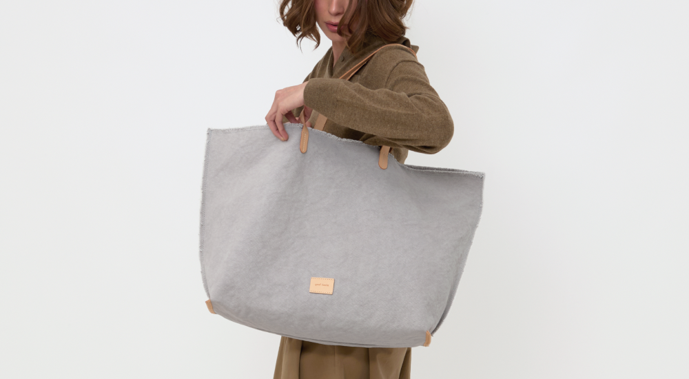 A woman in a brown outfit holds a large, light gray tote bag with tan handles against a plain white background.
