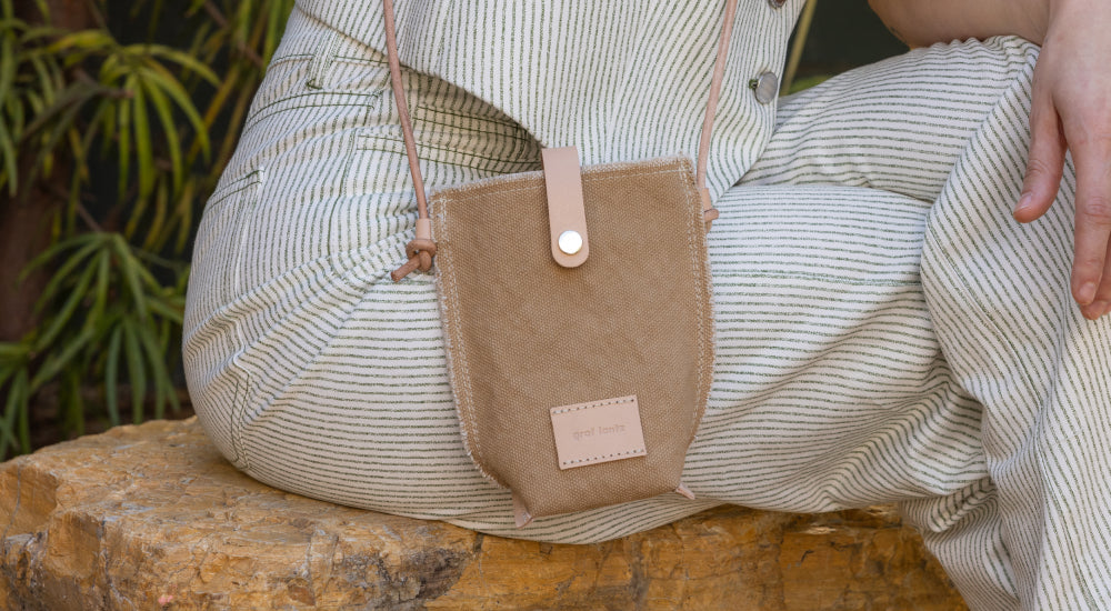 A person wearing striped light-colored pants sits on a rock, with a small tan crossbody bag resting on their knee. The bag has a buttoned flap and a small rectangular label.