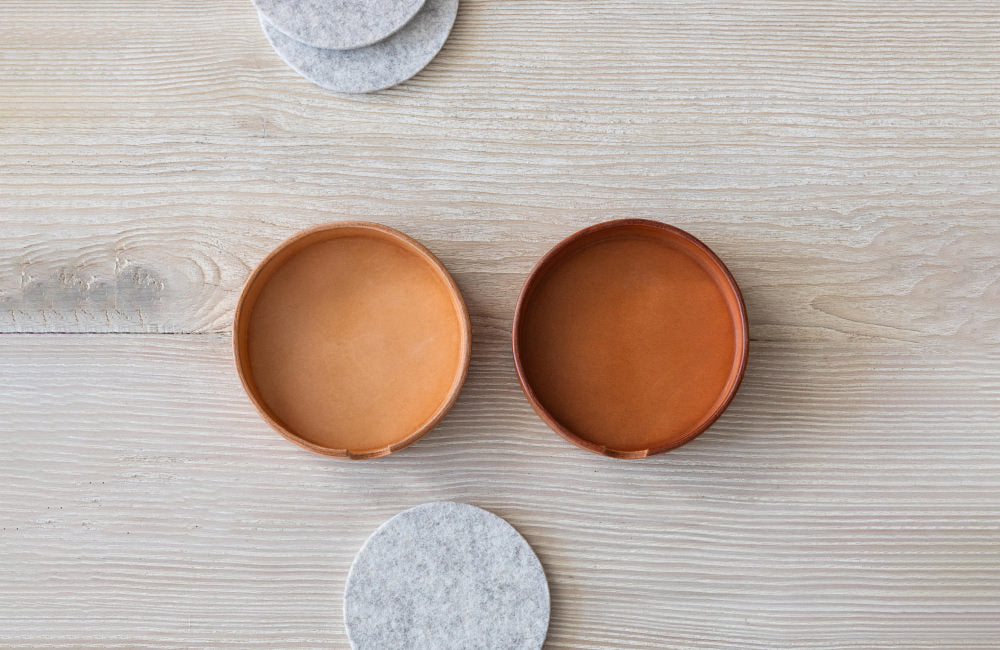 Two vachetta leather coaster holders, the left coaster holder is new while the other showcases patina. Gray Merino wool coasters next to the coaster holders on wooden tabletop.