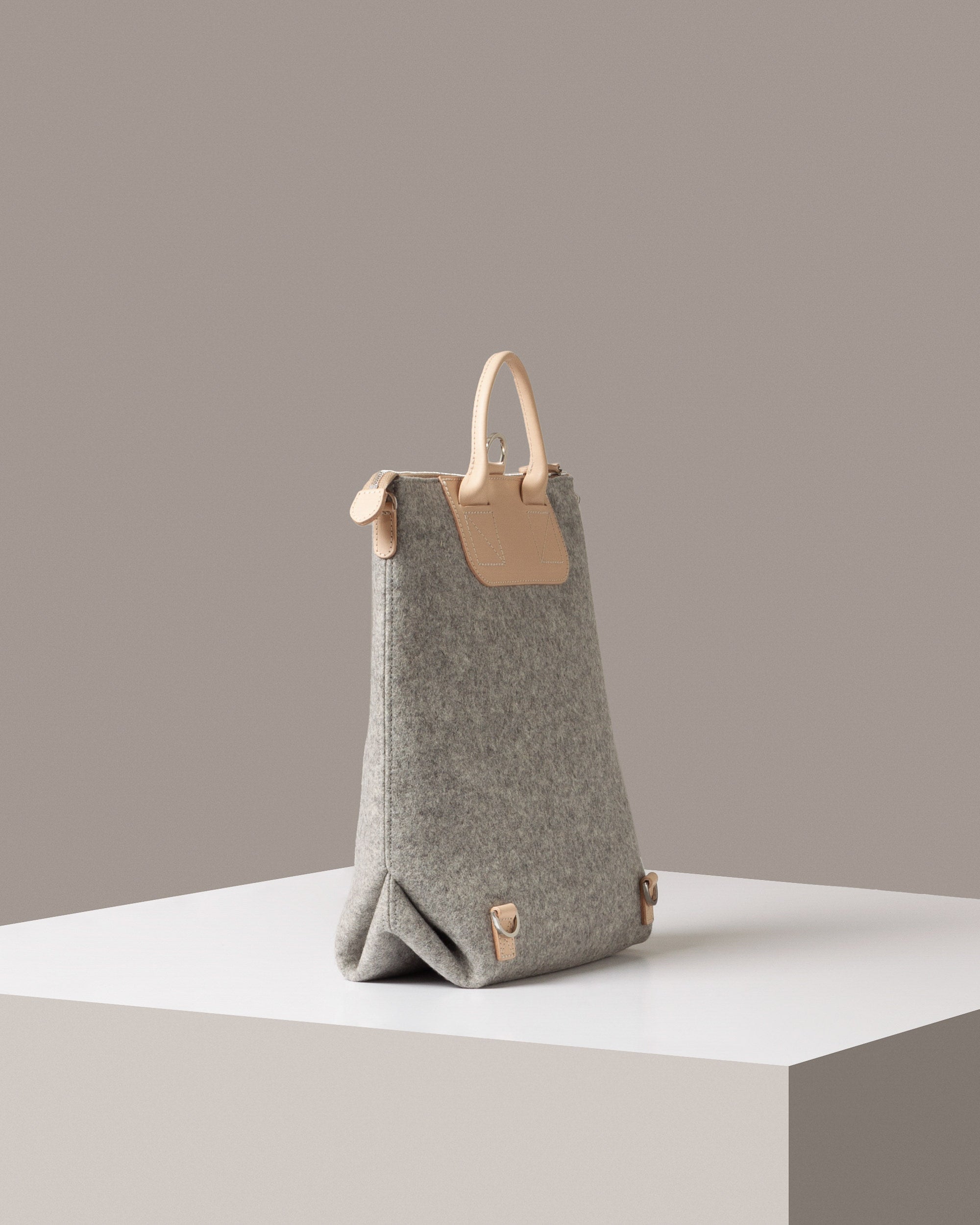A grey Bedford Merino Wool Felt Backpack on a white base is displayed in a side-view