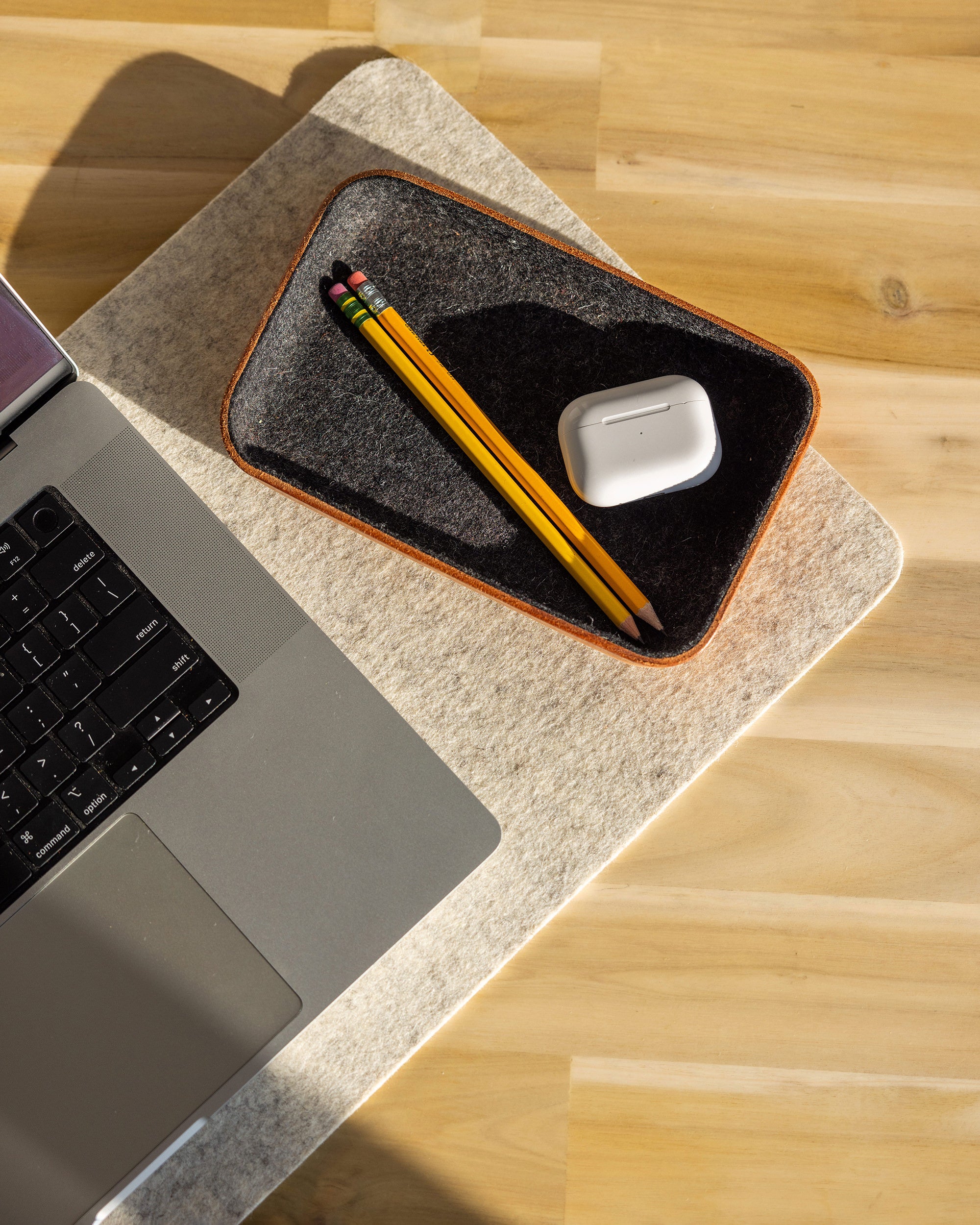 A laptop rests on a light wooden surface beside a graf lantz Merino wool catchall tray holding two yellow pencils and wireless earbuds in a white case, illuminated by natural sunlight.