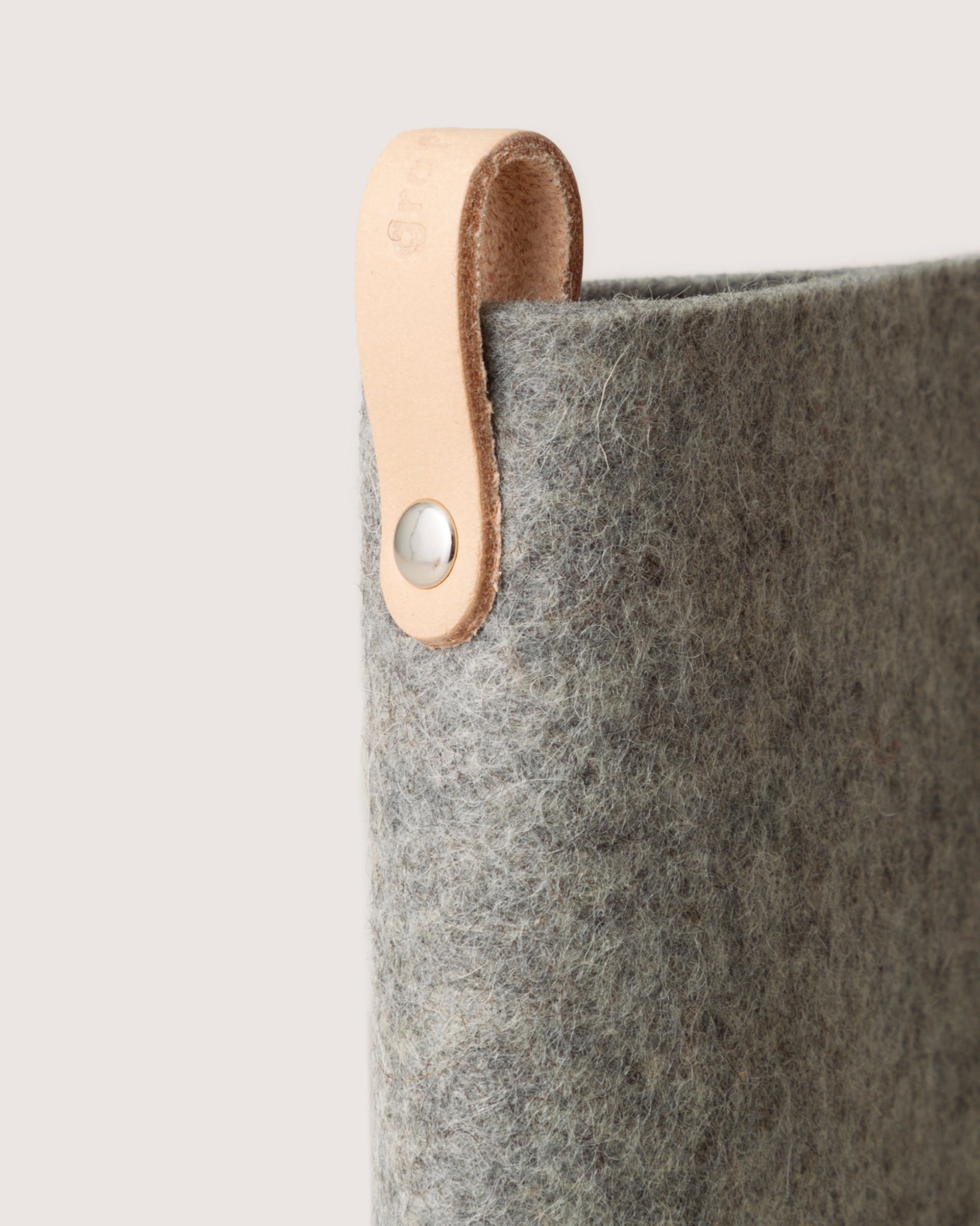 A close-up of a graf lantz gray felt bag highlights its textured Merino wool, accented with a light brown leather strap attached via metal rivet. The smooth leather finish contrasts elegantly against a neutral background, offering a stylish eyewear protection solution.