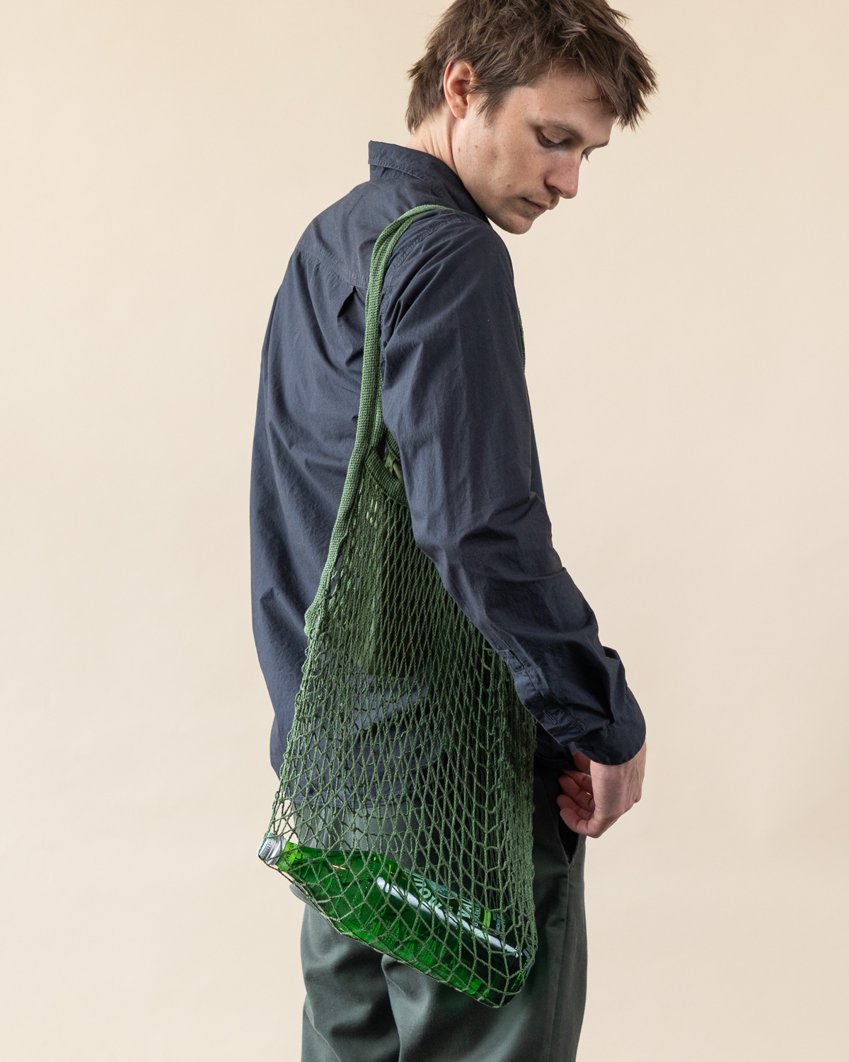 A person in a dark blue shirt and pants carries a Japanese fish market bag by graf lantz over their shoulder. The cotton net tote reveals three green glass bottles against a simple, light beige backdrop.