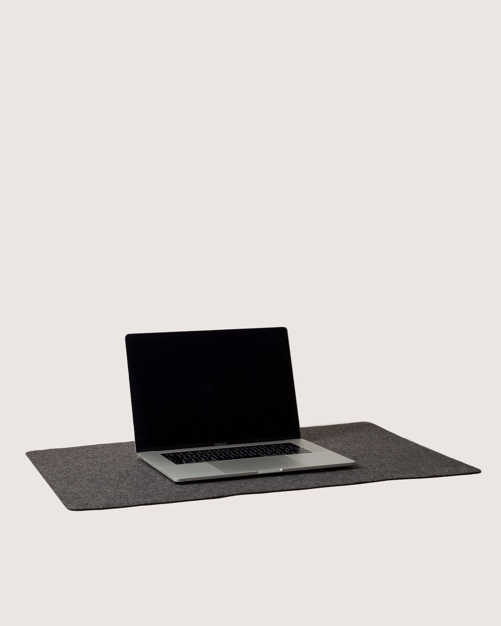 A gray laptop with a black screen sits on a graf lantz Mosen Desk Pad made of Merino Wool, set against a plain white background. The lid is partially open, revealing the keyboard and trackpad in this sleek workspace.