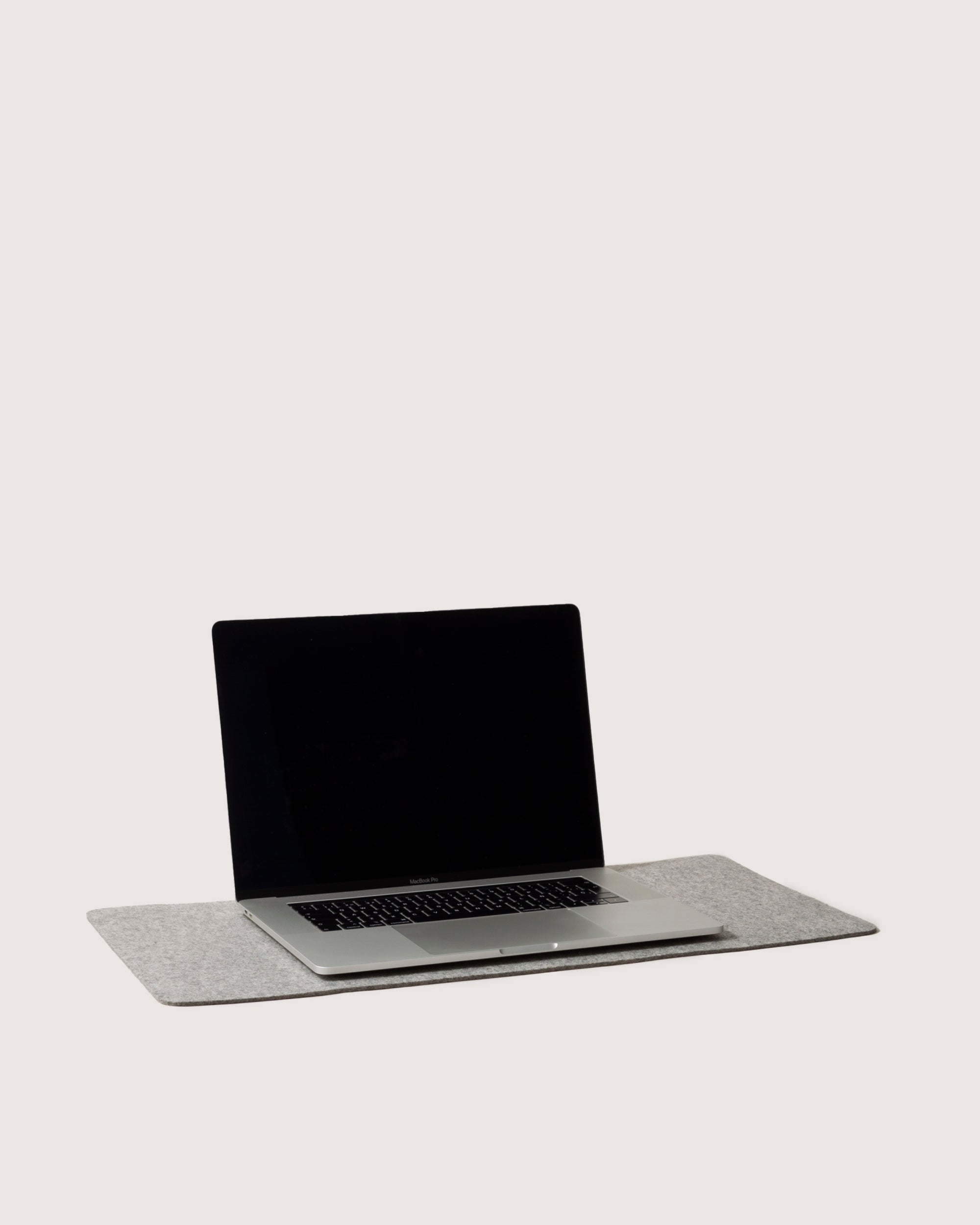 A sleek laptop rests open on a graf lantz desk pad crafted from light gray Merino wool, set against a plain white backdrop. The screen is blank, displaying its keyboard prominently, creating a minimalist and modern workspace aesthetic.