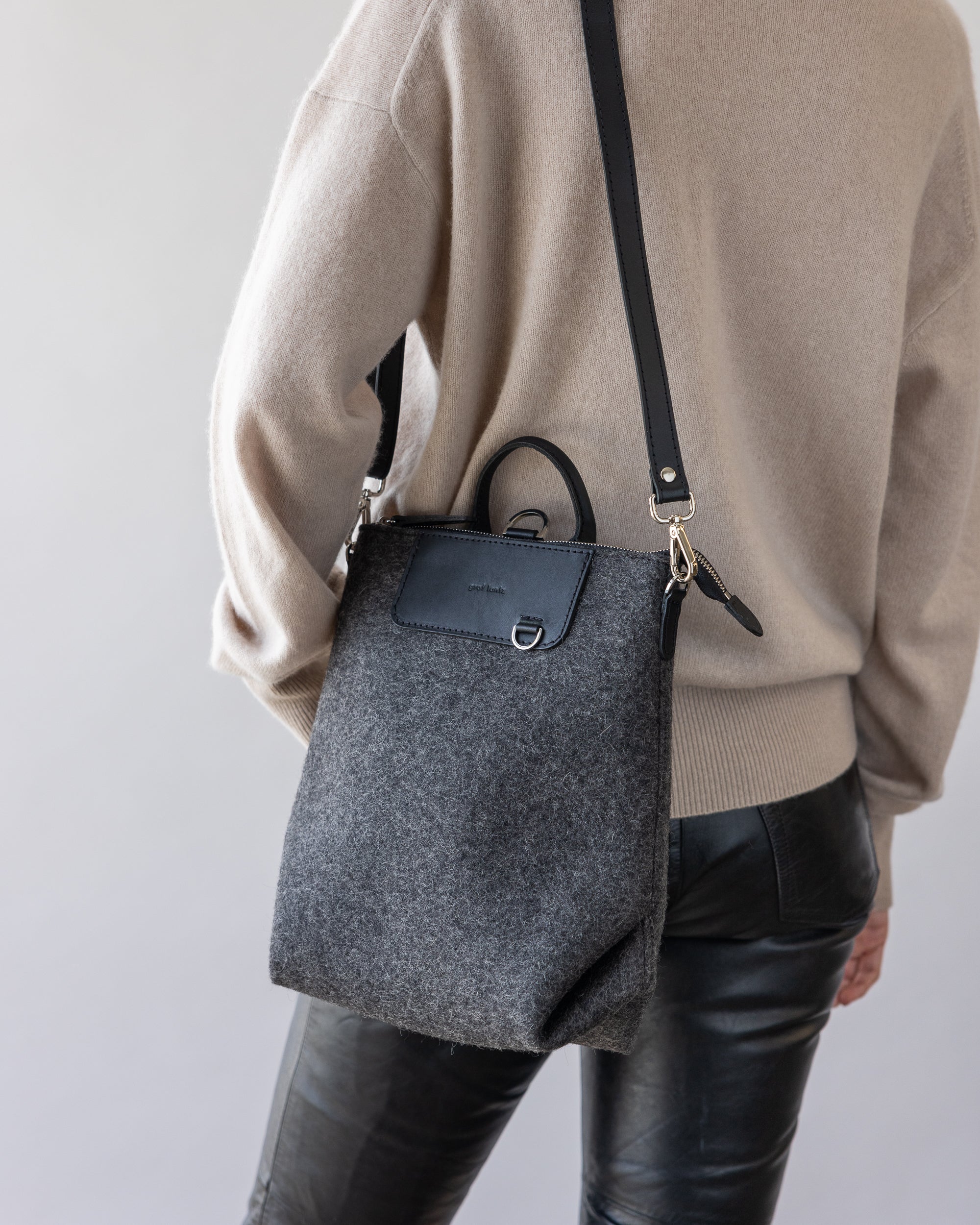 A person wears a beige sweater and black leather pants while carrying a graf lantz bag made from dark gray felt with a black leather strap and flap. This sustainable piece features minimal metal details, blending seamlessly into the neutral gray background.