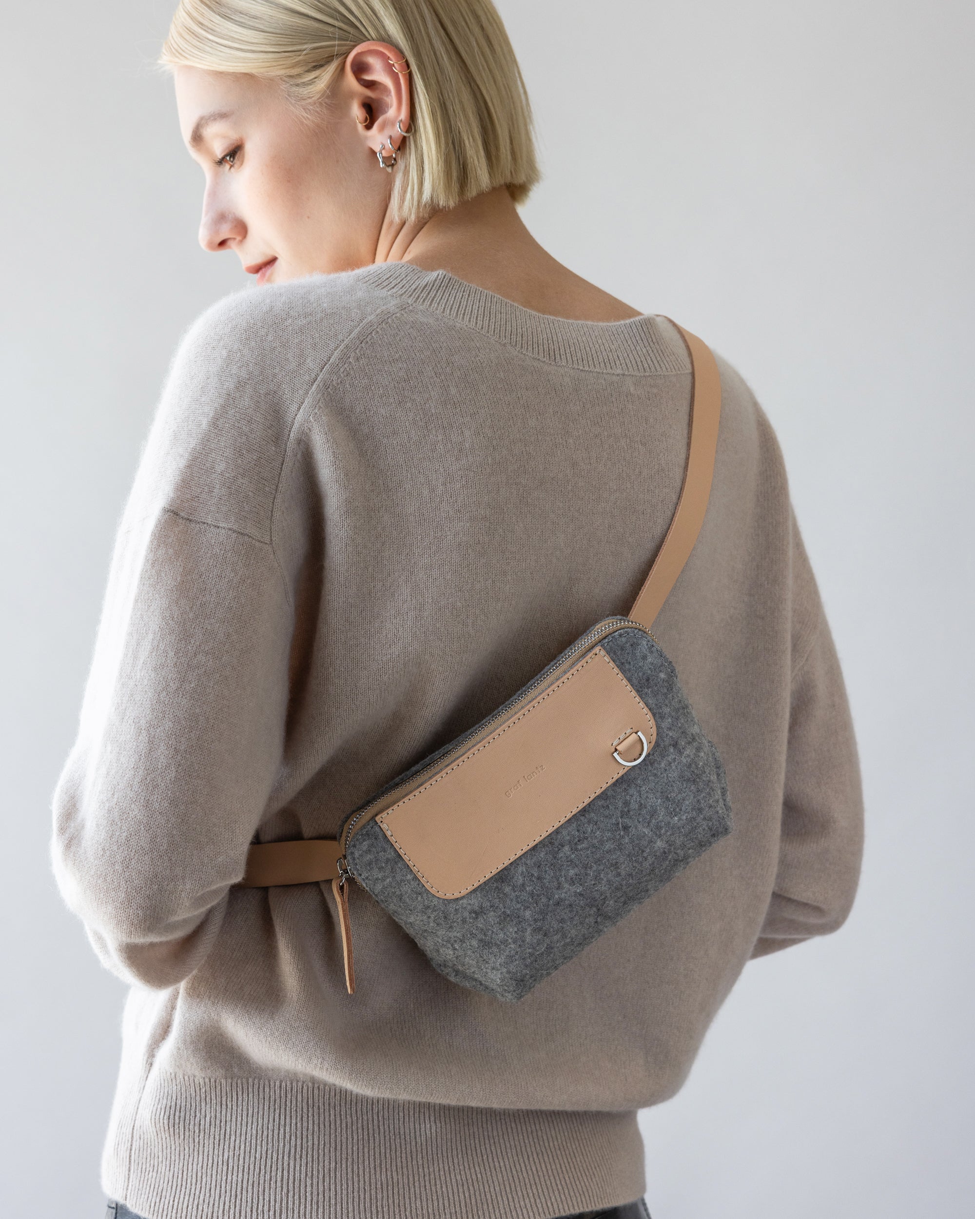 A person with short blonde hair wears a beige Merino wool sweater and a gray crossbody bag with beige accents by graf lantz, epitomizing hands-free convenience. They gaze sideways against a plain background.