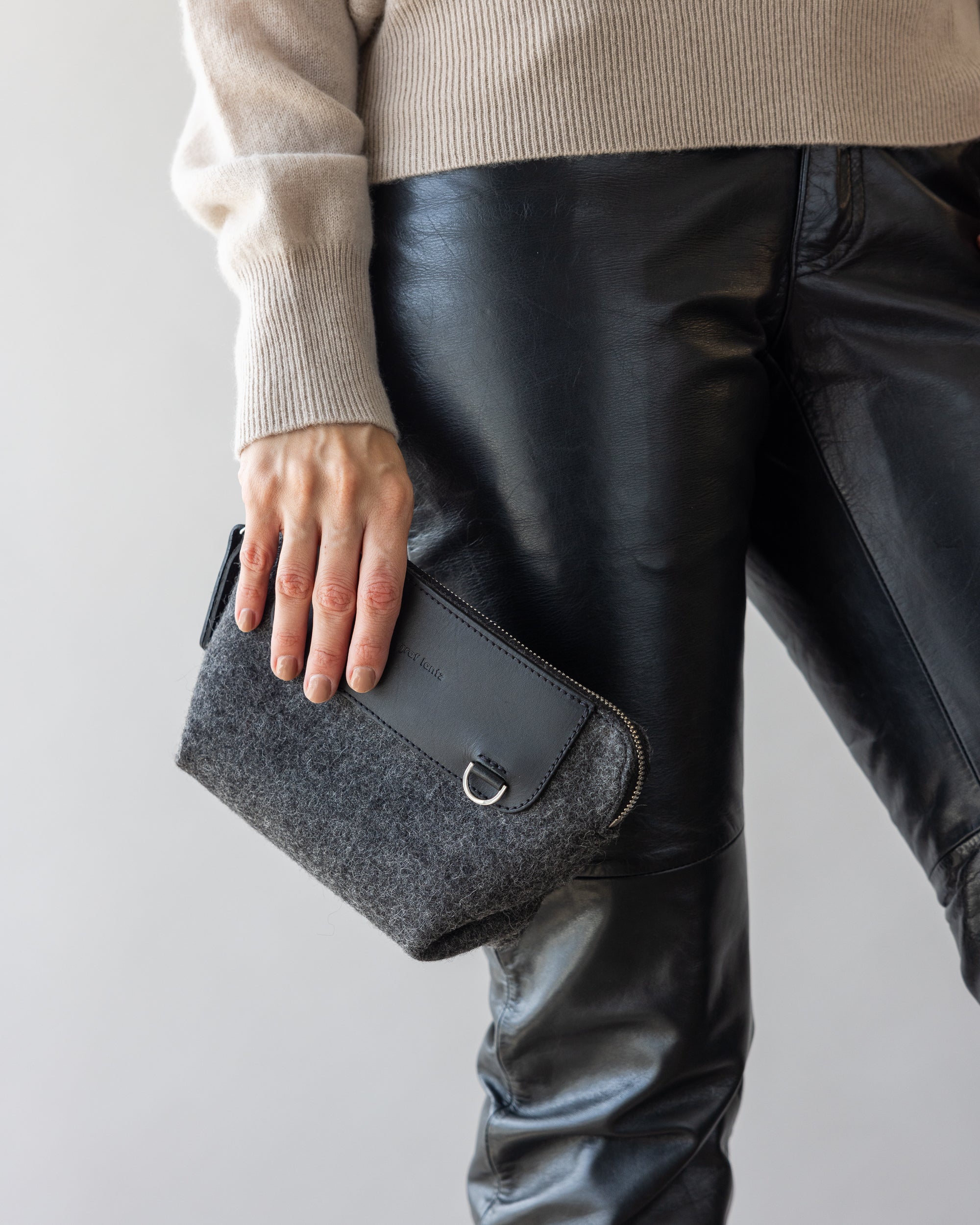 A person in black leather pants and a cozy merino wool sweater holds a sleek gray and black graf lantz clutch in their left hand, highlighting the stylish outfit and the clutch against a plain background.