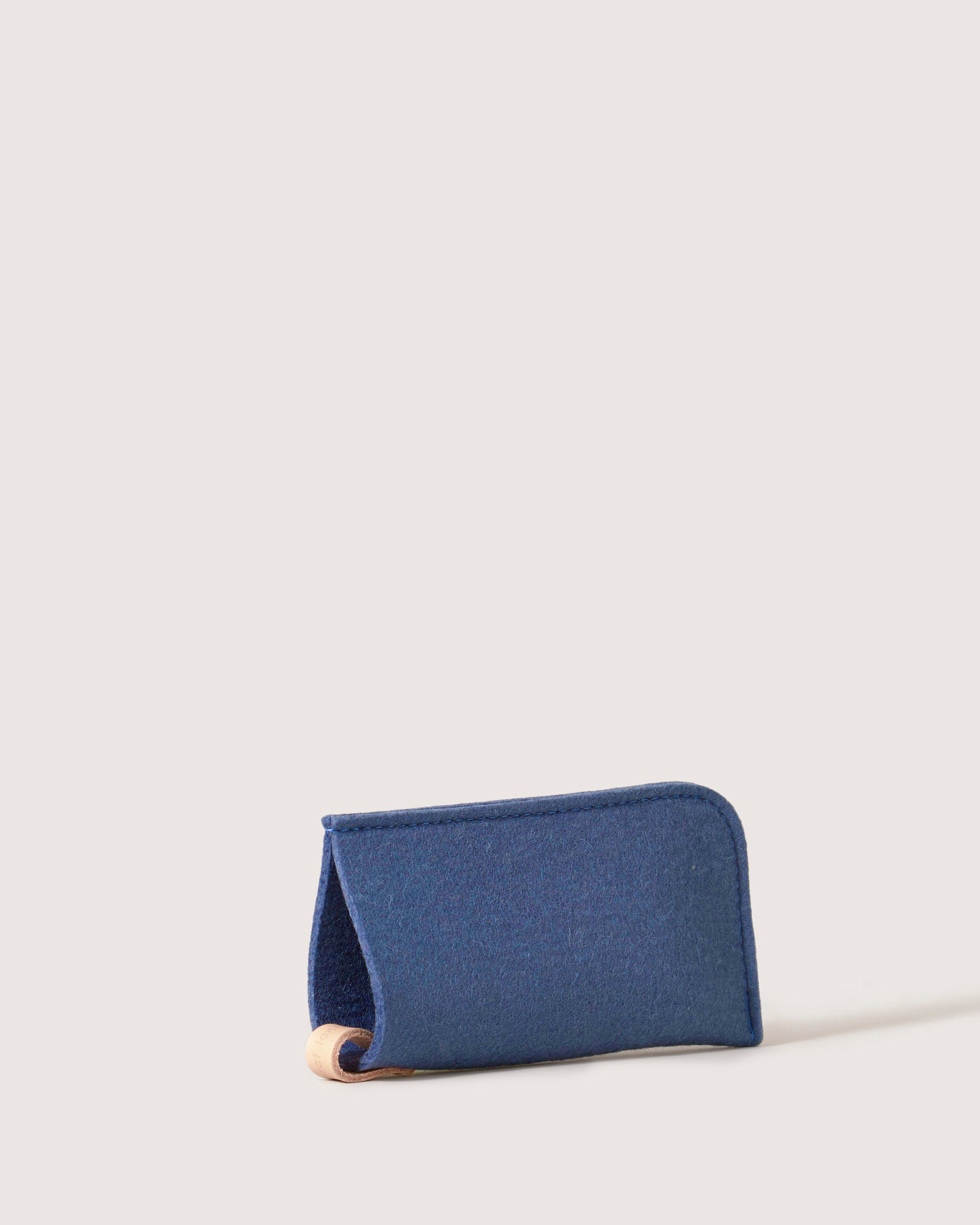 A blue fabric wallet by graf lantz rests on its side against a plain white background, subtly revealing an eyeglass sleeve with a wooden edge. The design is minimalist, combining style with utility.