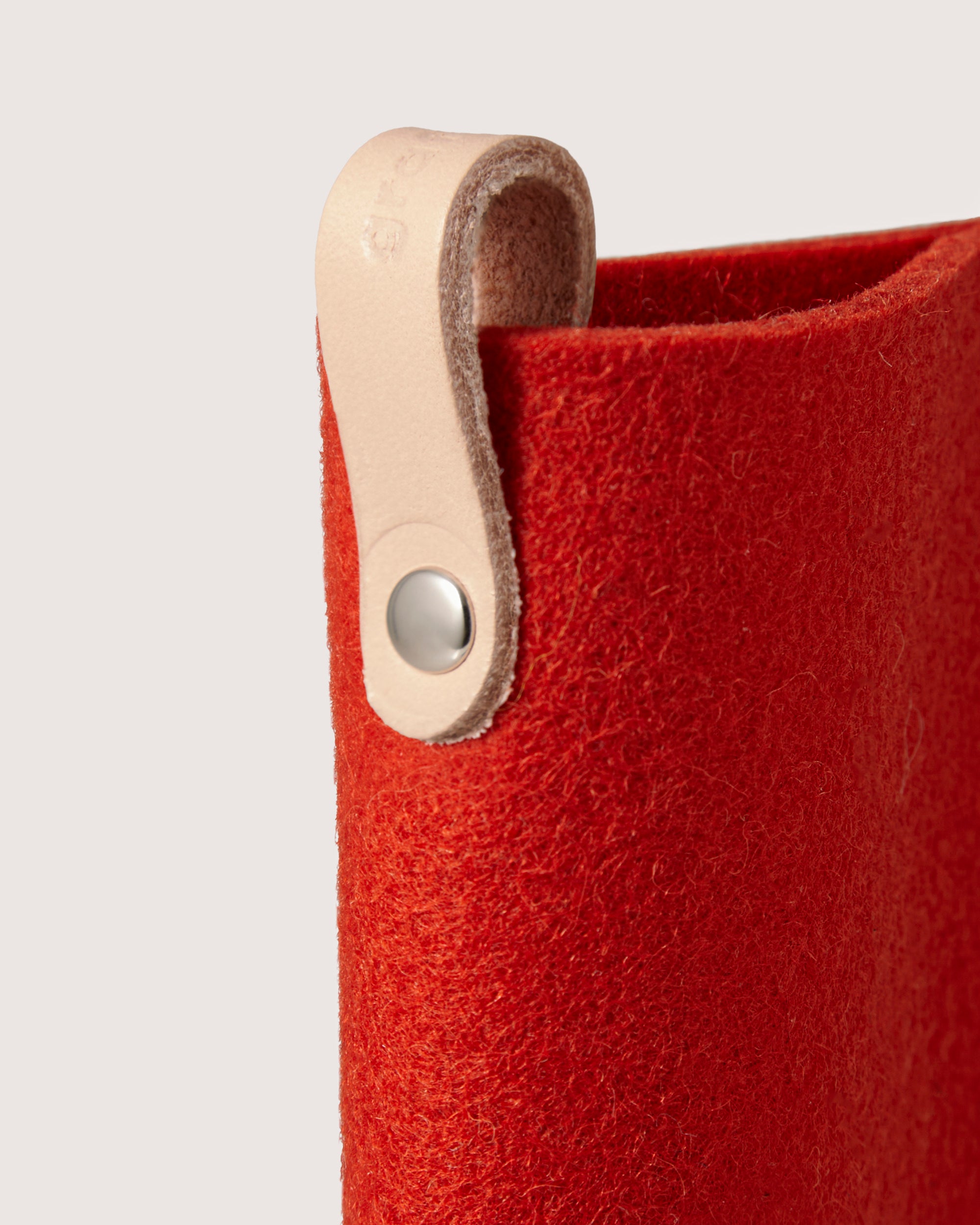A detailed view of the red felt bag by graf lantz, crafted from soft Merino wool, features a light tan leather strap secured with a silver button. The strap contrasts with the vibrant material and attaches to the top edge. Ideal for use as an eyeglass sleeve against a plain, light backdrop.