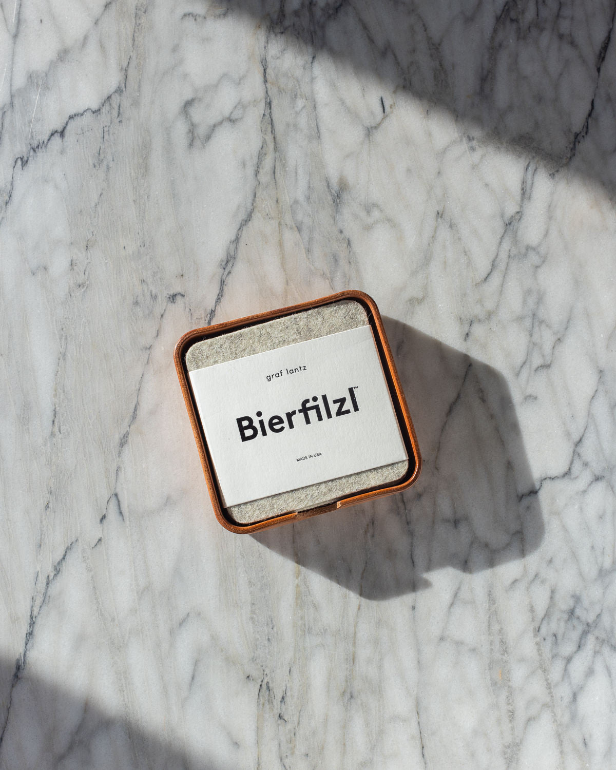 On a marble surface, square Merino wool coasters labeled Bierfilzl rest in a dish. The sunlight casts shadows, emphasizing their handcrafted quality by graf lantz in Los Angeles.