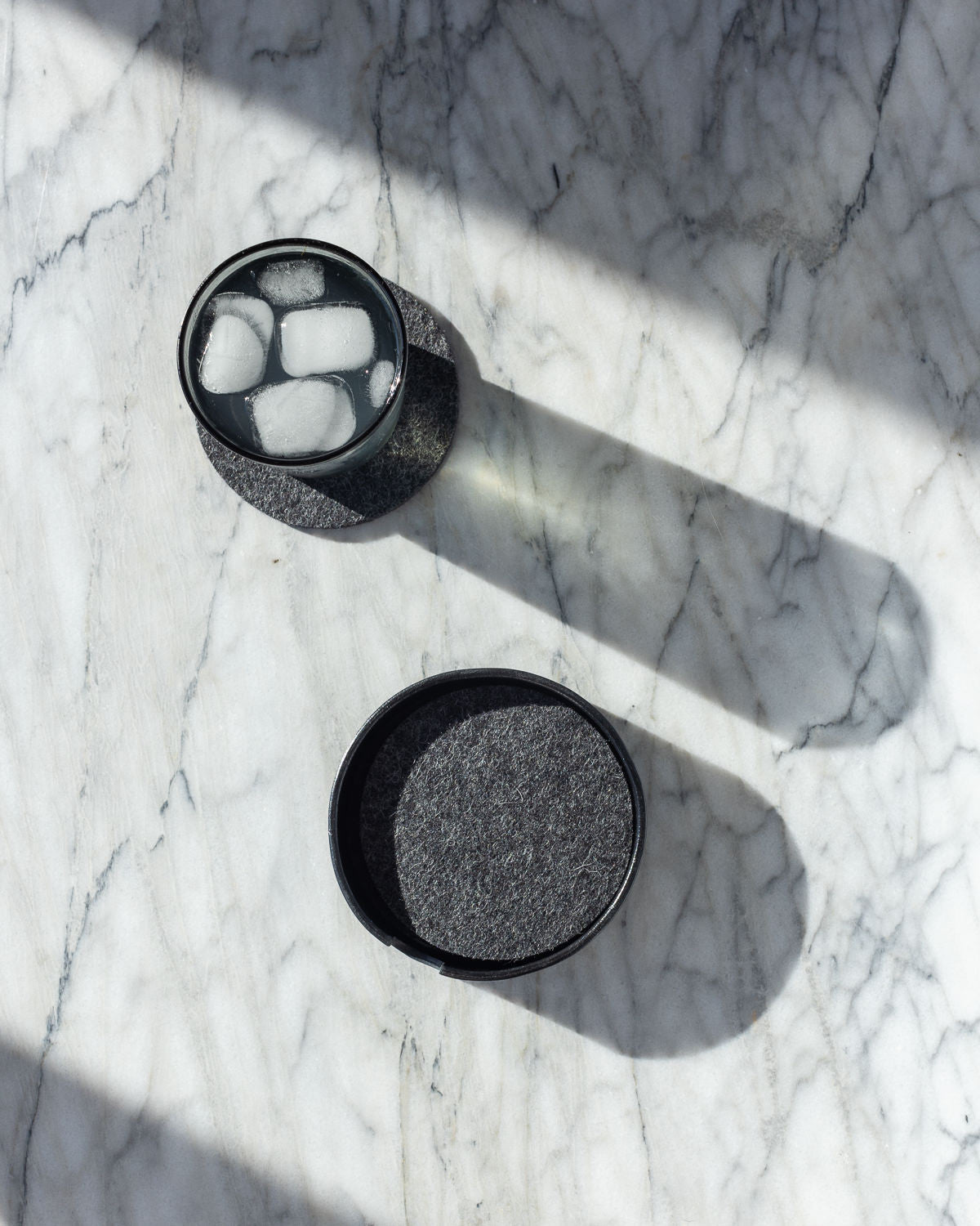A glass with ice cubes sits beside a closed container, both casting elongated shadows on a light marbled surface. Sunlight dances dramatically across the scene, reminiscent of the intricate craftsmanship found in fine graf lantz leather.