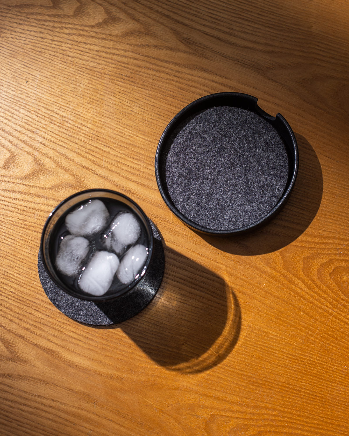 A glass of ice rests on a dark graf lantz Kobon leather coaster on a wooden table. Beside it, an open matching coaster holder from graf lantz adds to the scene. Sunlight highlights the wood’s texture and the artisanal craftsmanship of the items.