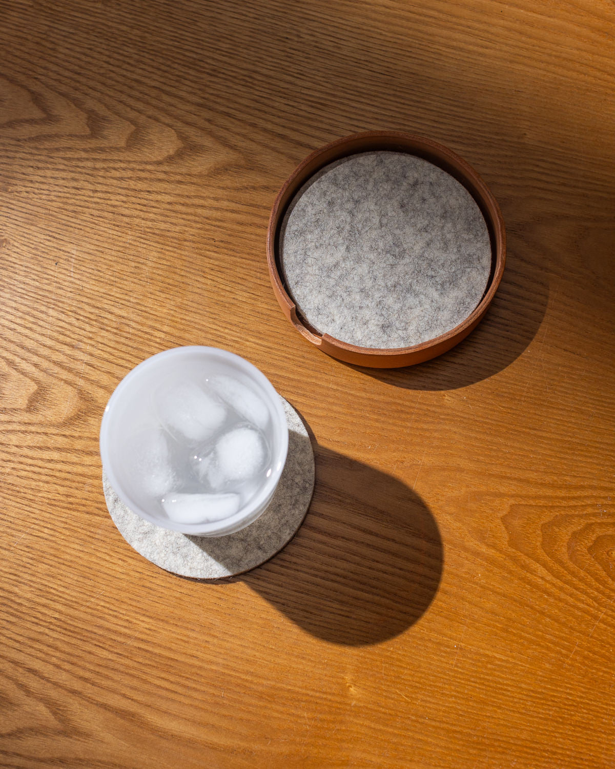 A white glass with ice cubes sits on a textured Merino wool coaster by graf lantz, placed on a wooden table. Nearby, a brown container with lid rests in a Kobon leather tray. Natural sunlight casts soft shadows over the artisanal scene handcrafted in Los Angeles.