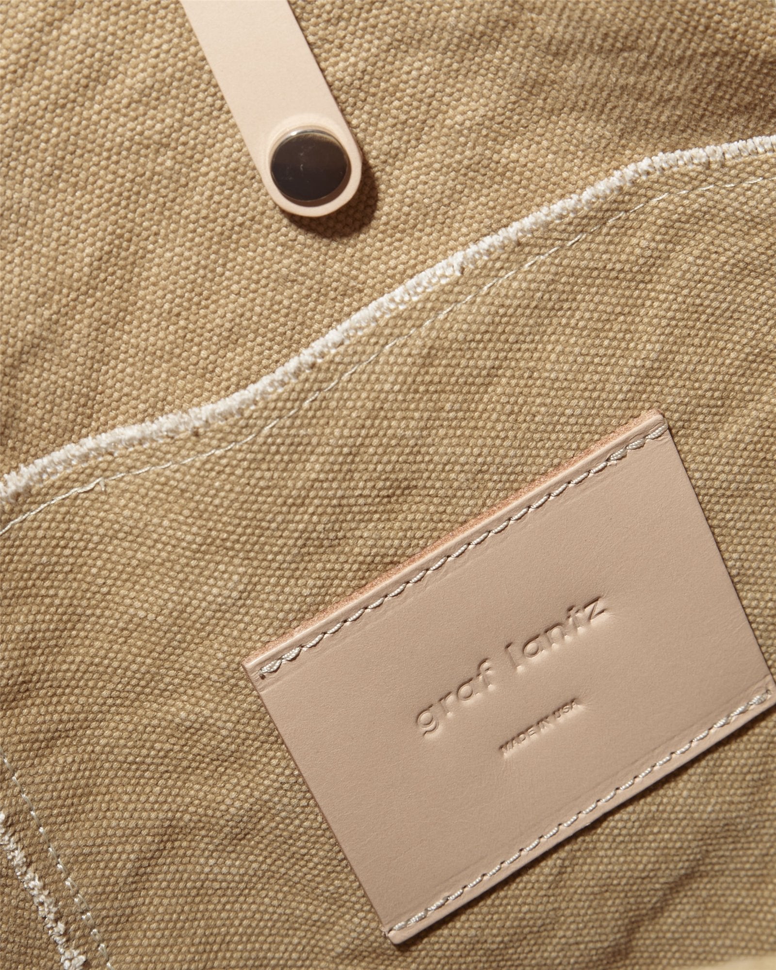 A close-up shows a tan, textured fabric bag with a stitched pocket and an embossed Graf Lantz leather tag. The durable cotton canvas tote has a fabric strap secured by a button and elegant leather handles.