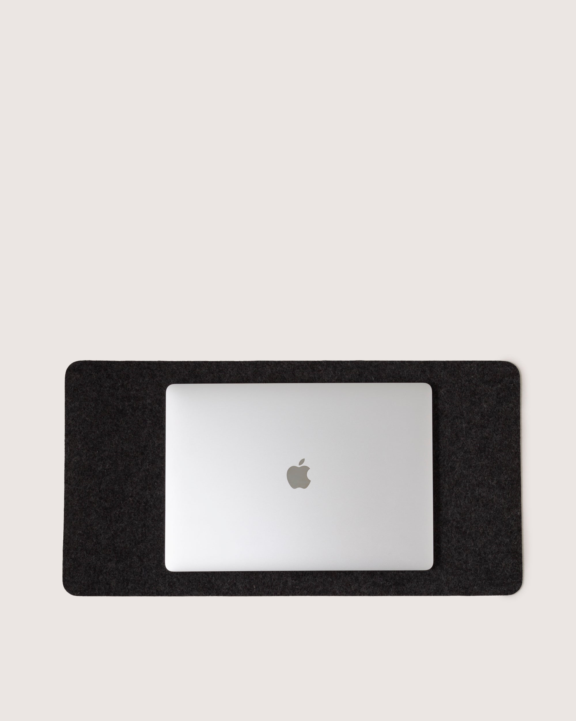 A silver laptop with a partially visible logo is on the elegant graf lantz Mosen Desk Pad, made from fine Merino Wool. With rounded corners, this pad contrasts elegantly with the plain light background. Closed, the laptop shows off its smooth metallic surface in a chic workspace.
