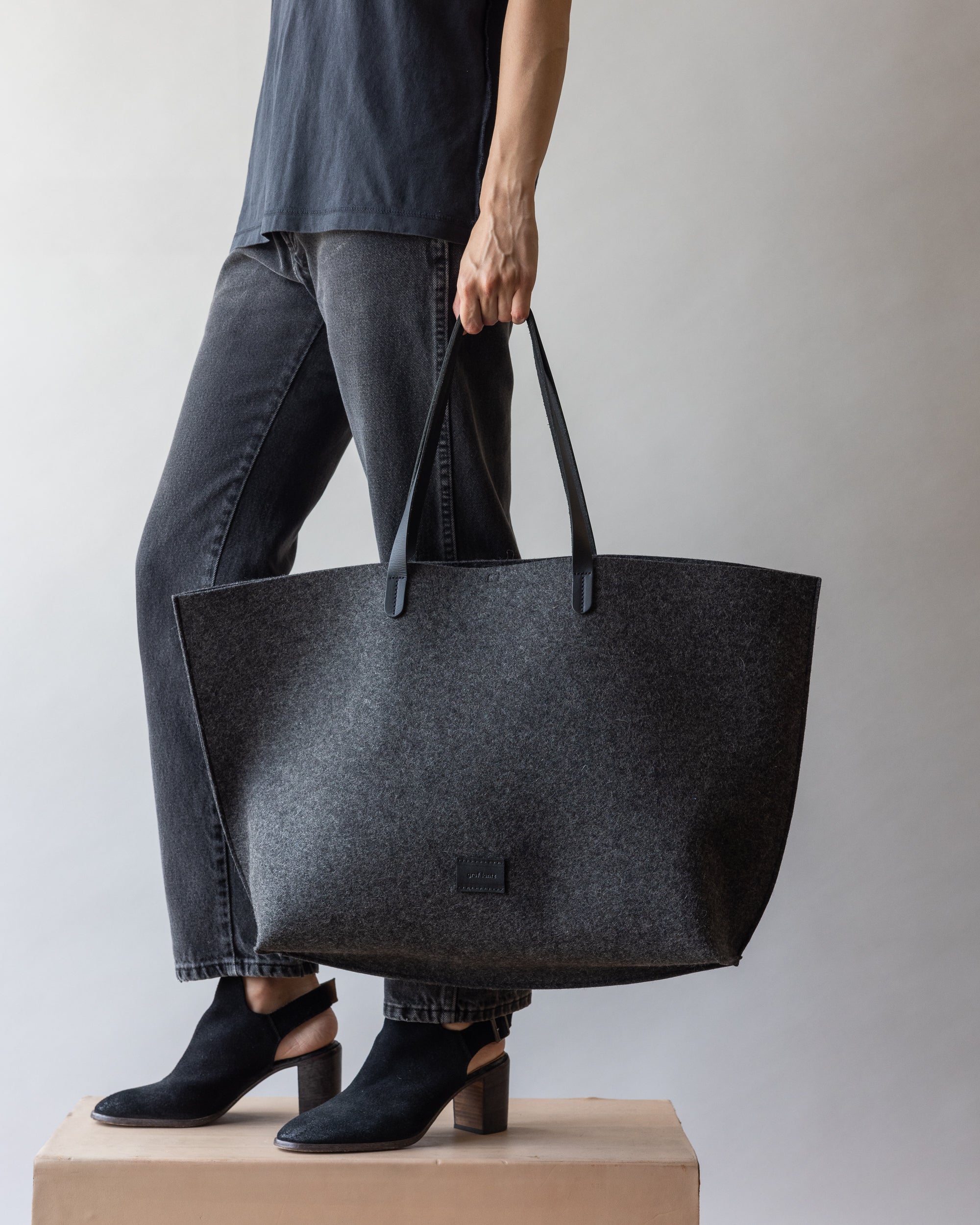 On a wooden platform, a person holds a large gray tote bag featuring sleek leather handles by graf lantz. Theyre wearing a dark shirt, gray jeans, and black ankle boots against a simple light backdrop that enhances their sustainable style.