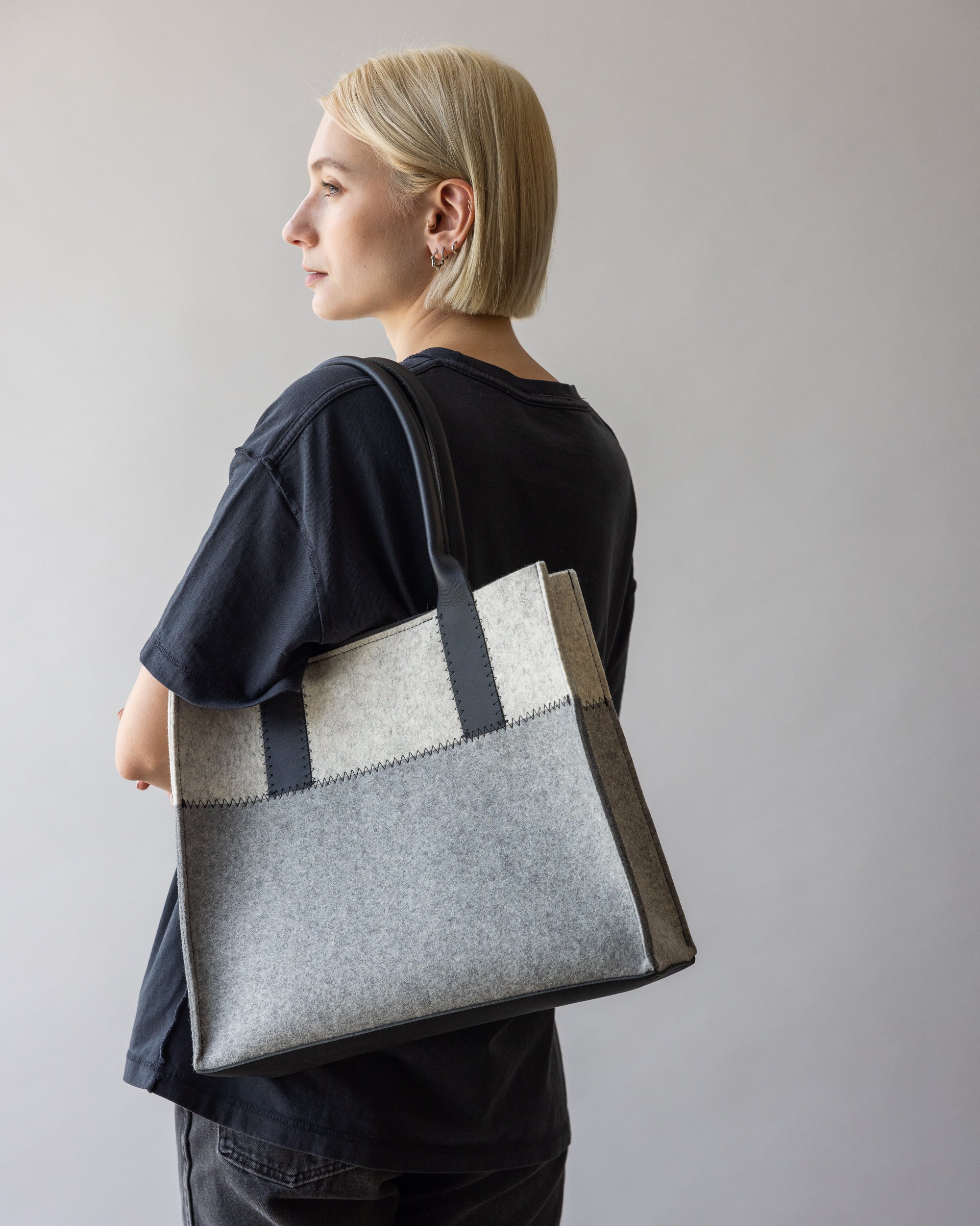 A person with short blonde hair stands sideways, wearing a loose black t-shirt and holding a graf lantz Jaunt Midi Tote in two-tone grey with black handles. The mid-size carryall exudes elegance under the neutral, soft lighting against the plain background.