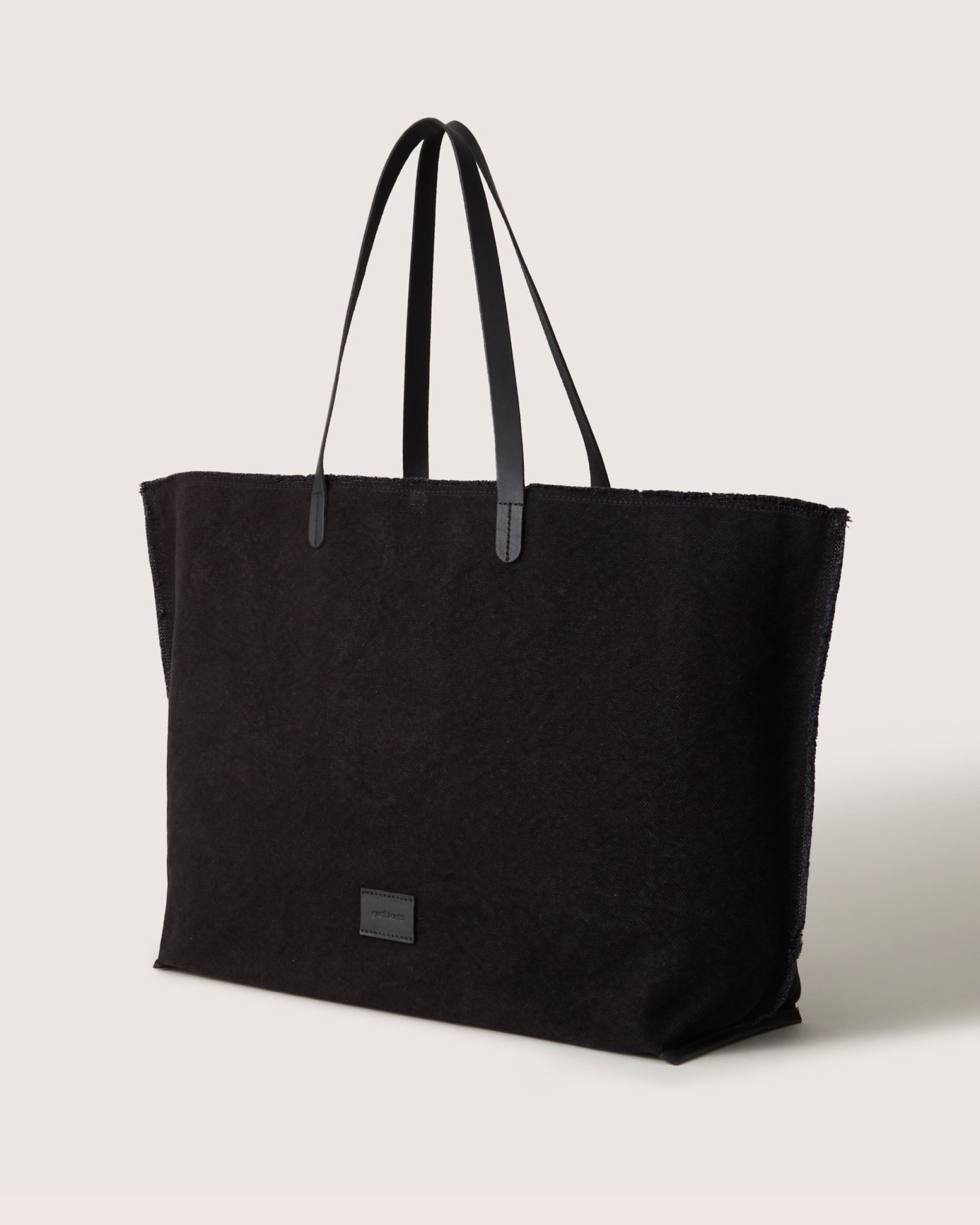 The large Hana Canvas Boat Bag by graf lantz features a simple, black tote design with two thin genuine leather handles and a small rectangular label on the front. Its design includes pockets for easy organization, all set against a plain white background.