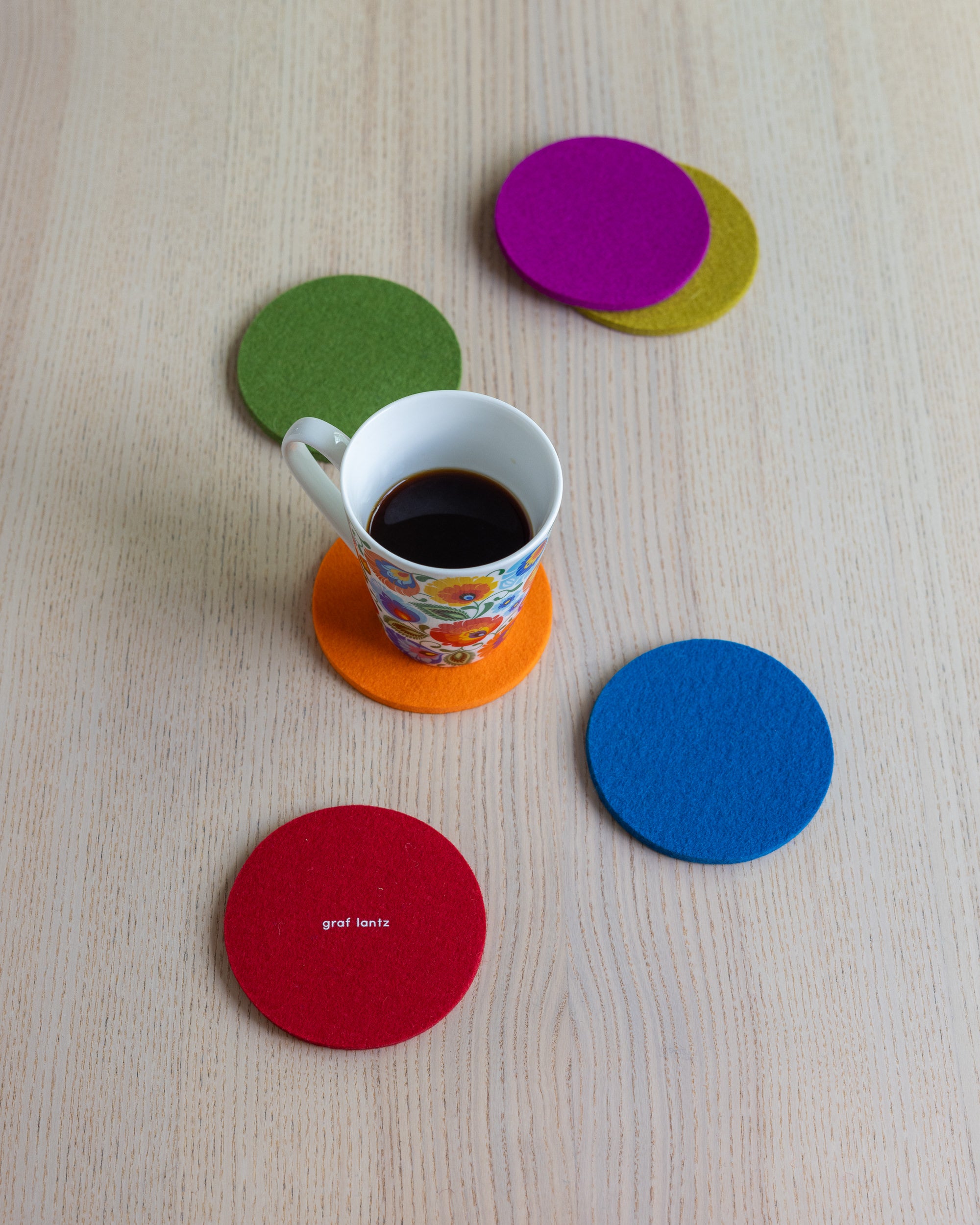 A white mug with colorful patterns sits on an orange coaster atop a light wooden table, surrounded by four graf lantz round coasters in green, yellow, purple, and blue. The set includes a red German Merino wool coaster adorned with white text.