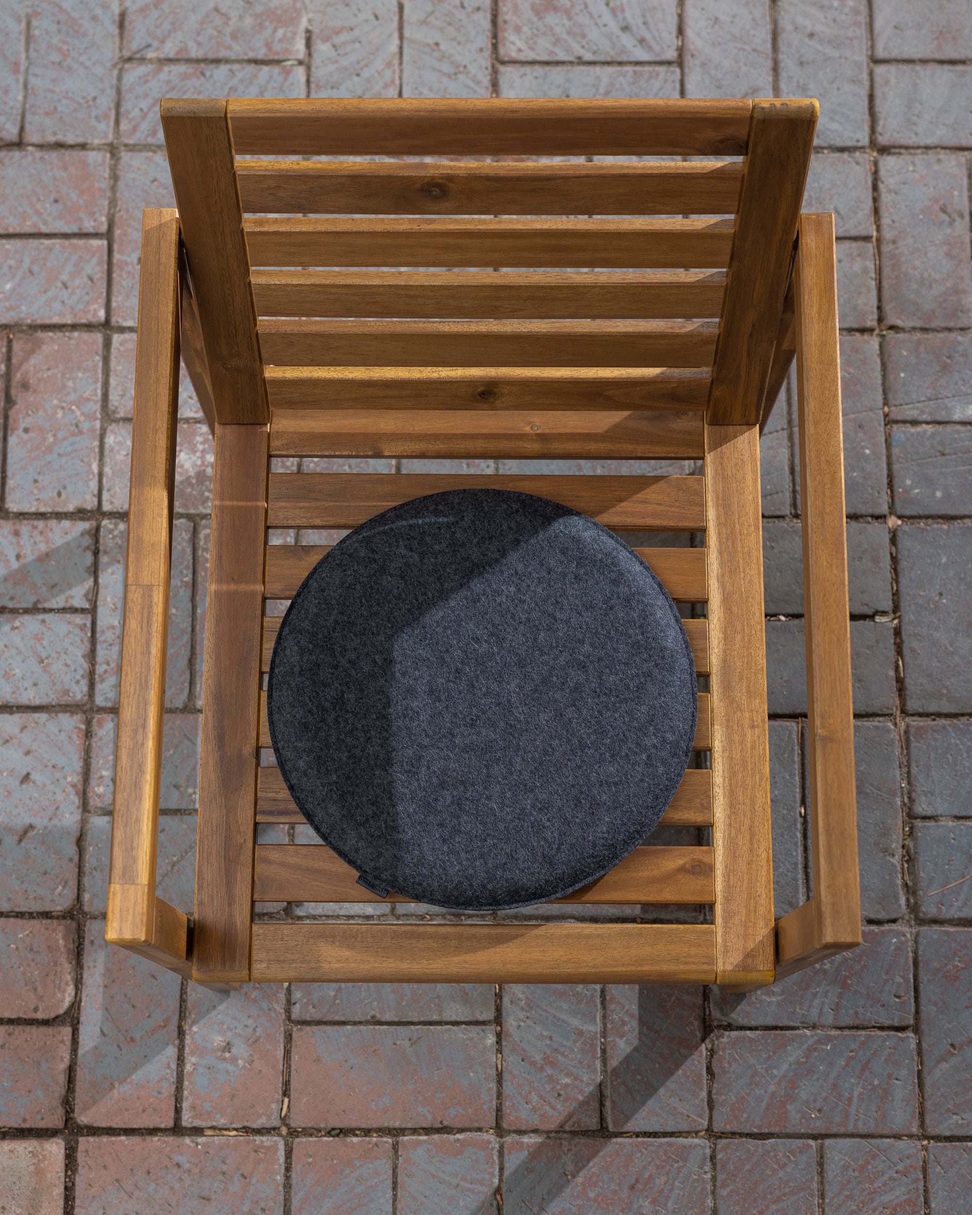 Top view of a wooden chair featuring a graf lantz Merino wool round, dark gray cushion on the seat. The chair rests on a tiled patio with varied brown and gray shades, creating a tranquil scene perfect for meditation. Shadows stretch gracefully across the area.