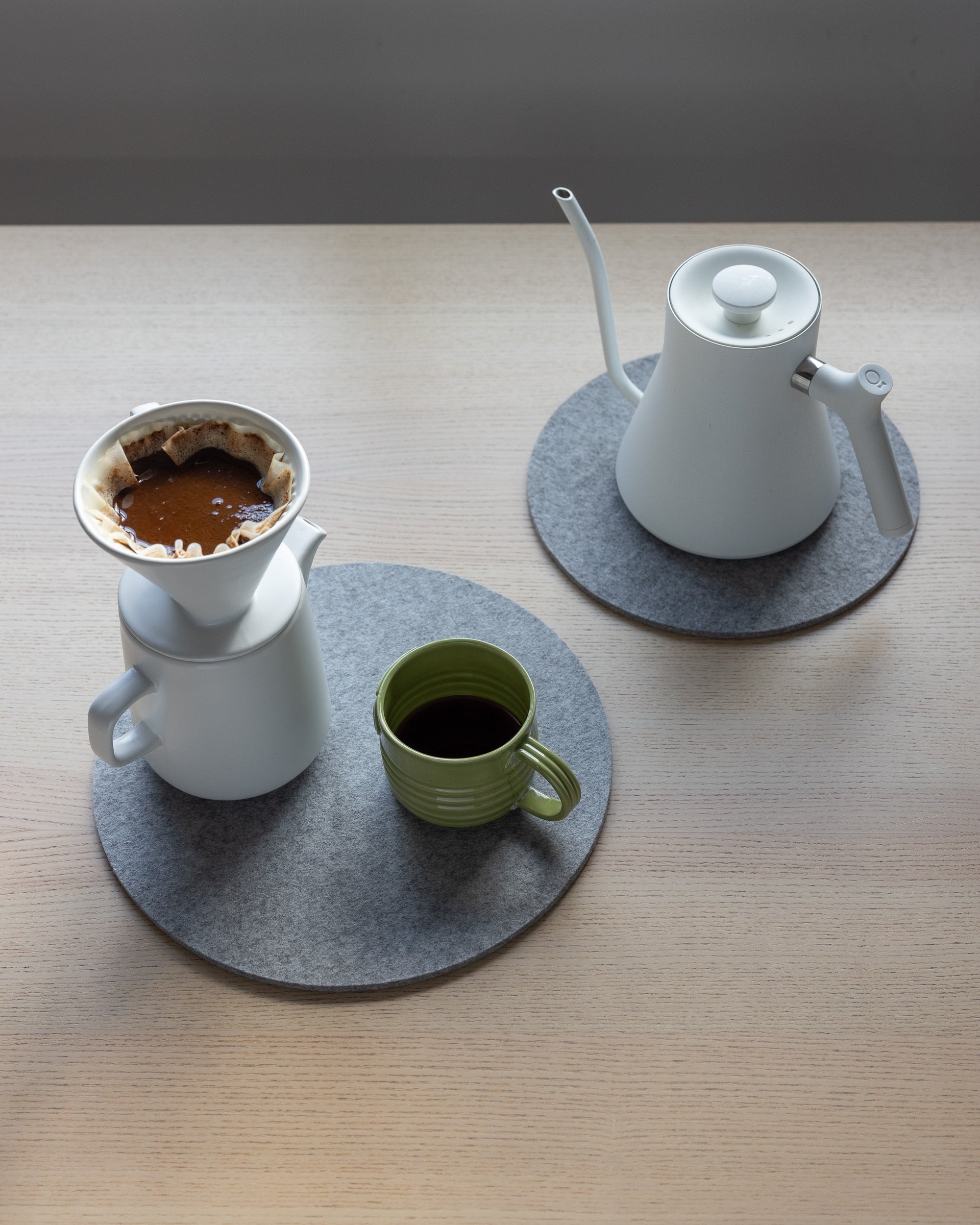 A light wooden table holds a pour-over setup featuring a white ceramic dripper and carafe. A green mug sits on a graf lantz gray moisture-wicking felt mat, while a white gooseneck kettle rests on an identical heat-resistant graf lantz mat.