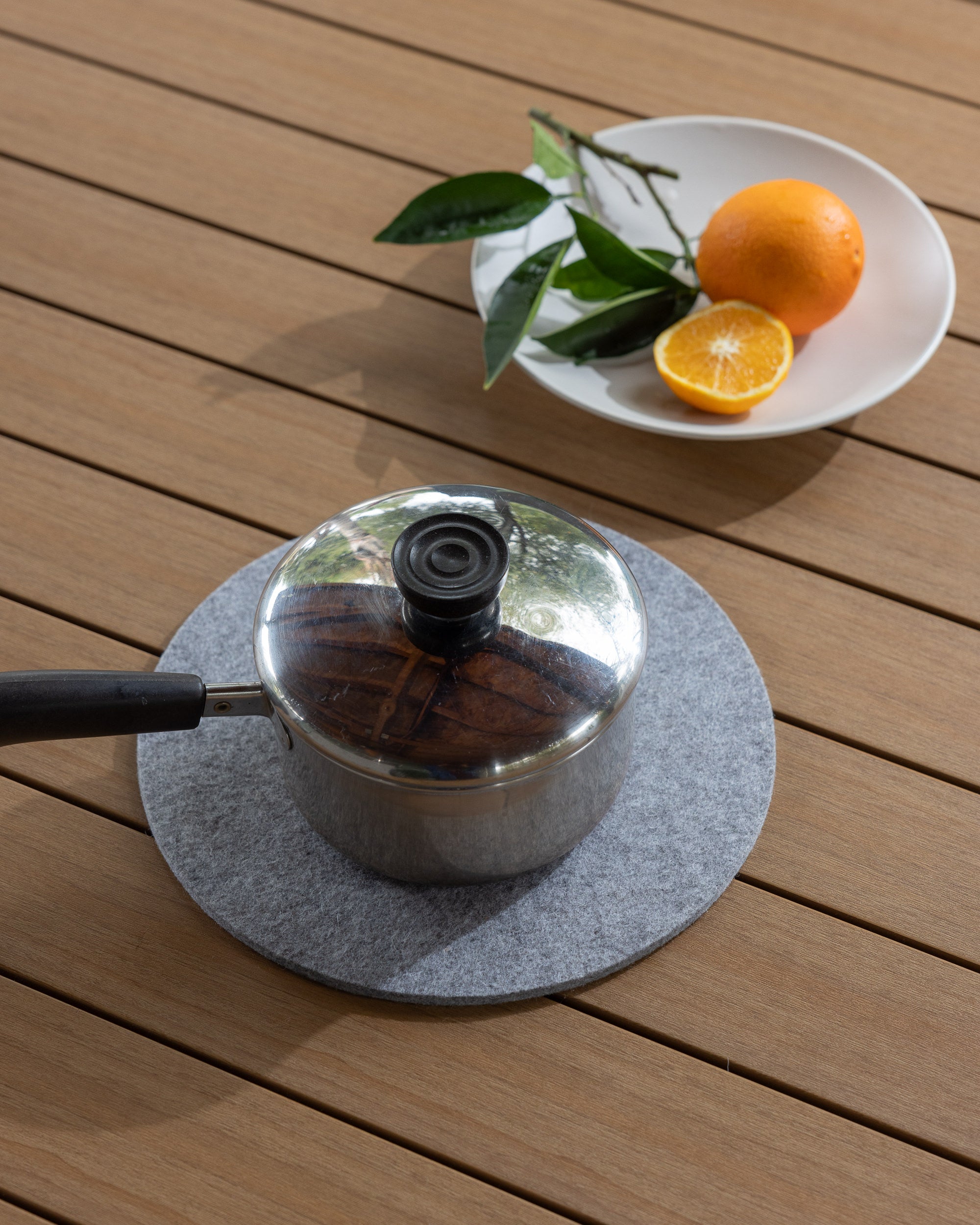 A small metal saucepan with a black handle sits on a graf lantz gray felt mat crafted from moisture-wicking Merino wool, placed on a wooden surface. Beside it is a white plate holding an orange, a halved orange, and some green leaves.