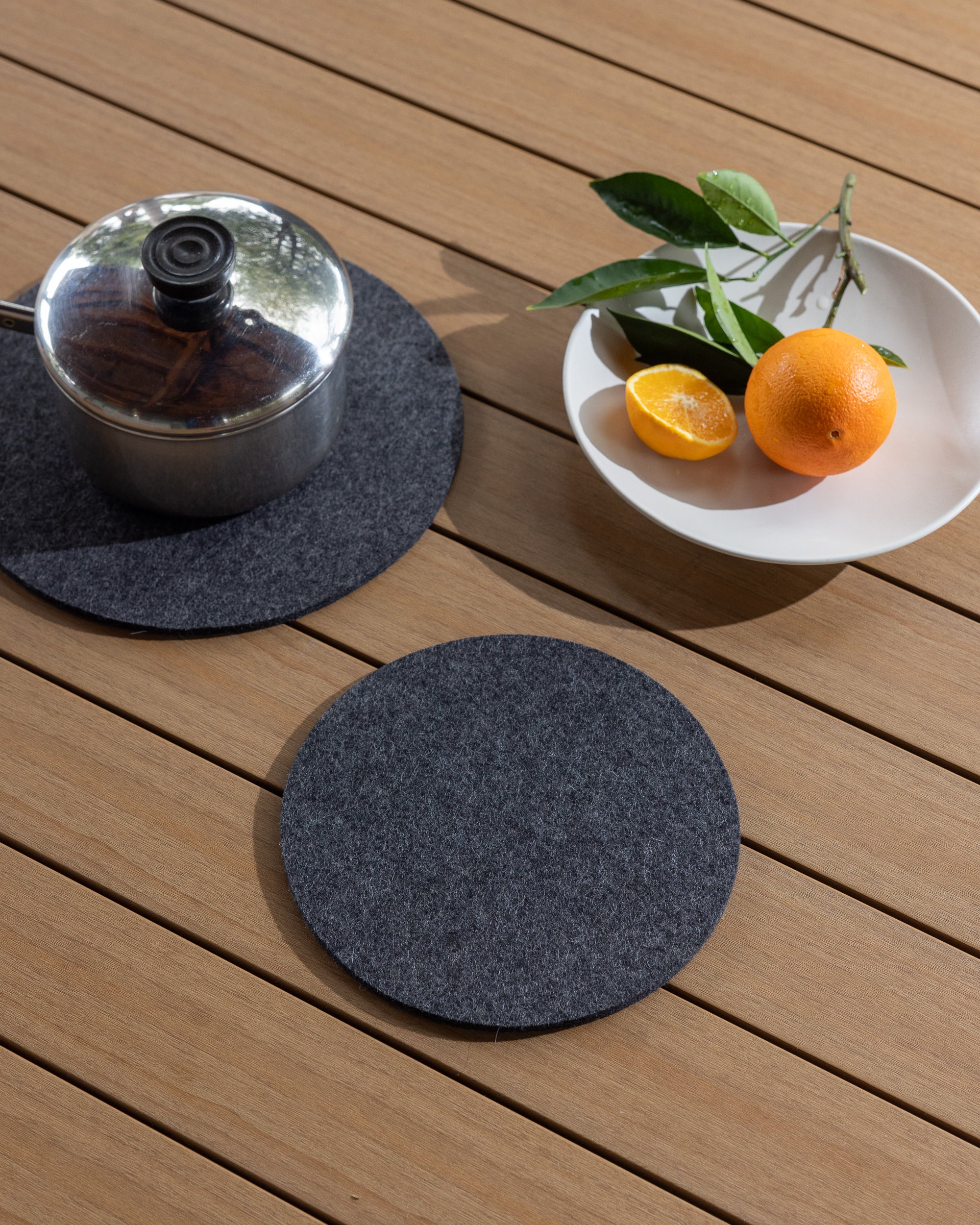 A metal pot with a dark lid rests on a round gray Merino wool mat by graf lantz, highlighting its heat-resistant quality on a wooden surface. Nearby, a white bowl holds an orange, half an orange, and green leaves. An identical trivet is placed in front.