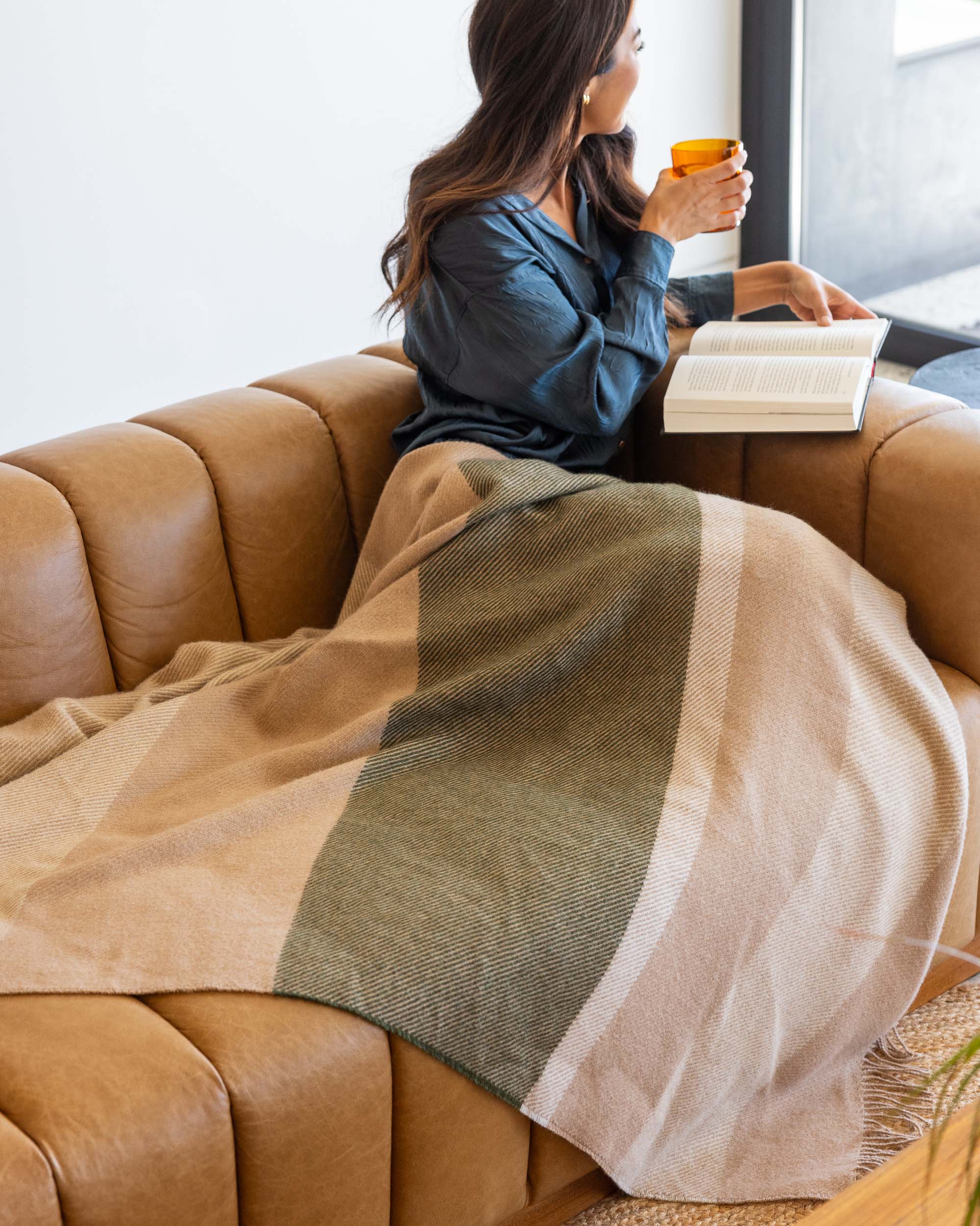 A woman sits on a brown leather sofa, wrapped in a beige and green plaid oversized alpaca blanket. She holds an orange graf lantz mug and gazes out the window, with an open book on her lap. The room is well-lit and cozy.