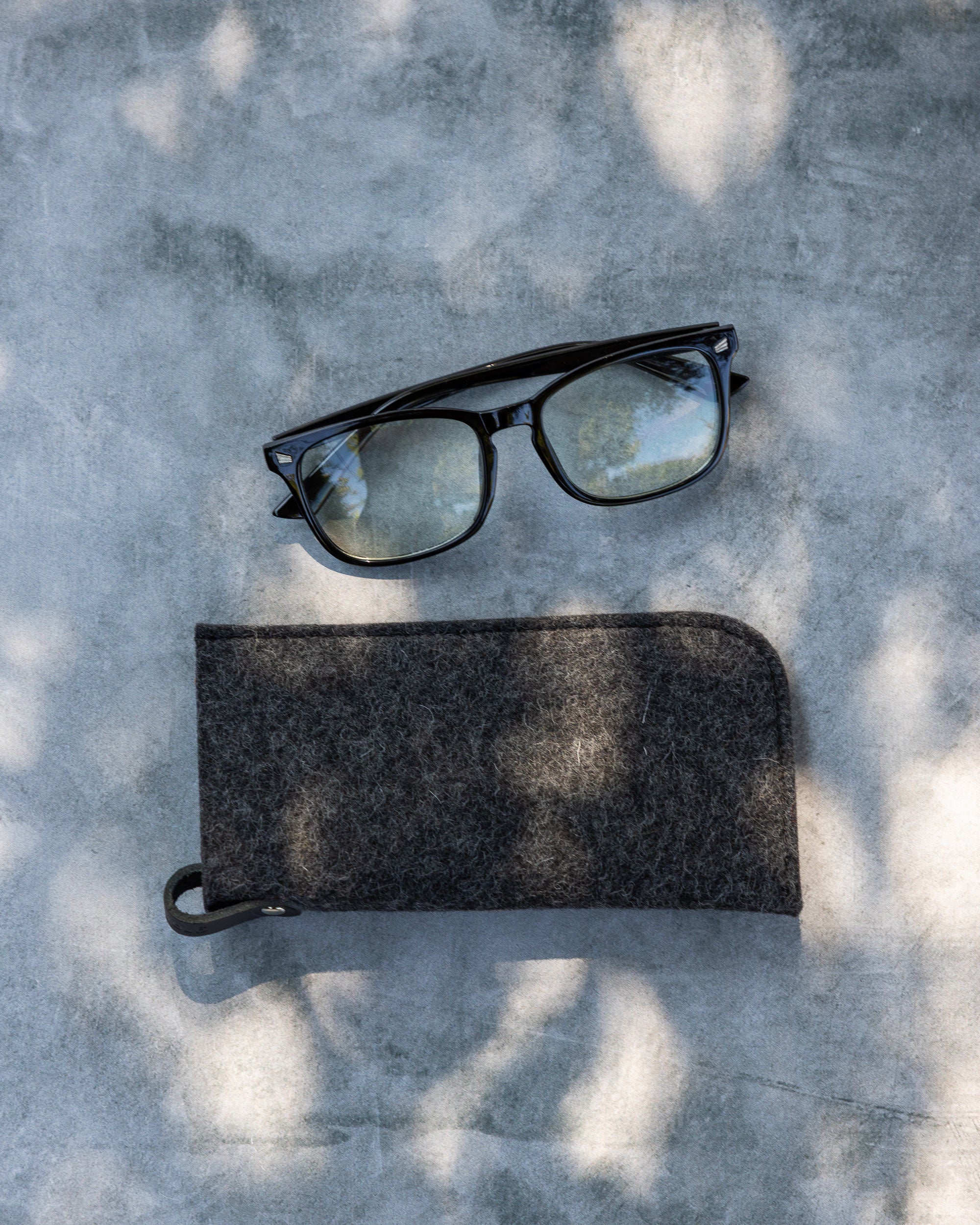Black rectangular eyeglasses with clear lenses sit on concrete in dappled sunlight. Below them is a gray felt eyeglass sleeve from graf lantz, designed for optimal protection, featuring a zipper and small hook.