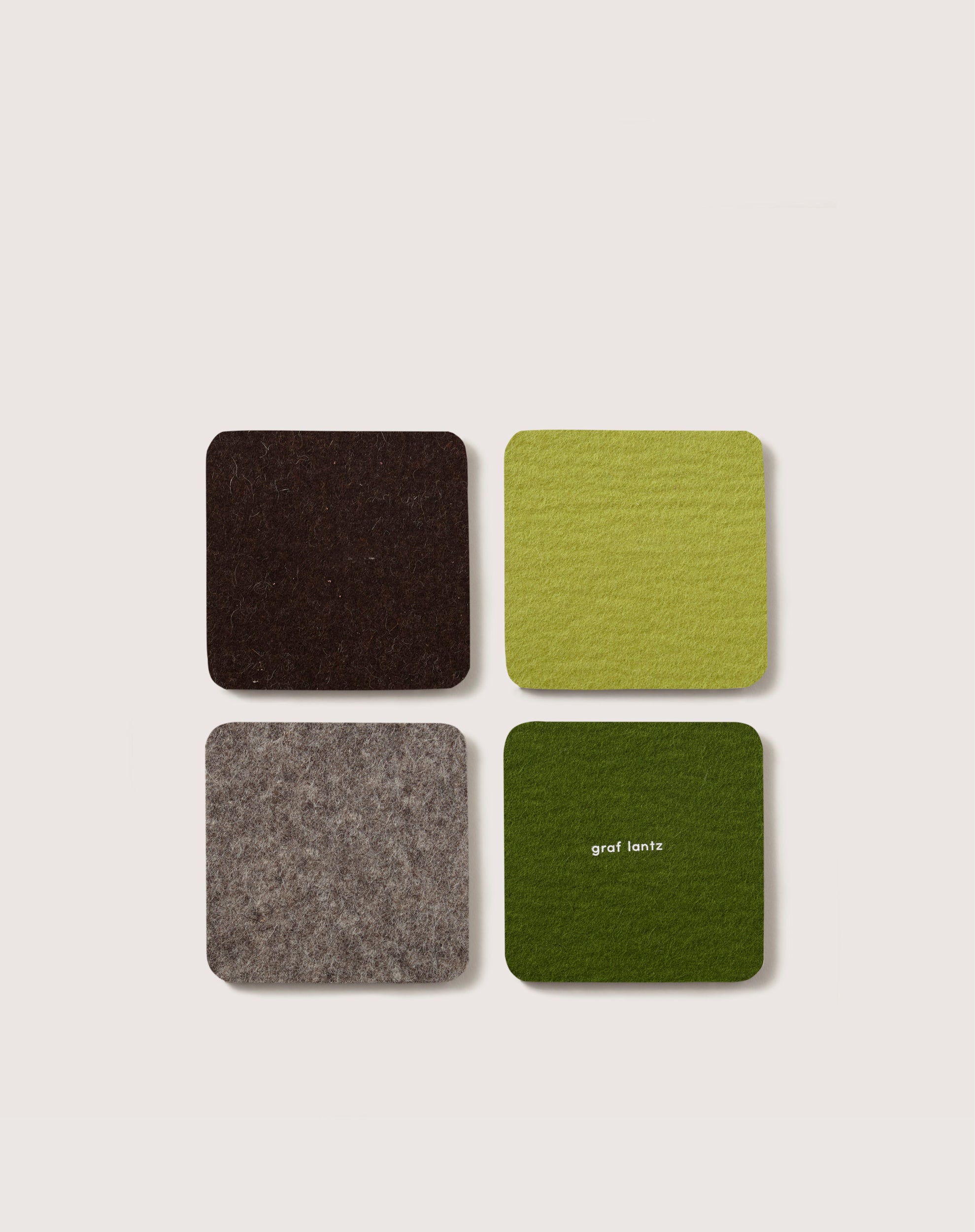 A set of four square coasters, made from premium Merino wool, features dark brown, light green, gray, and dark green hues. The dark green coaster is elegantly embossed with graf lantz, adding a touch of German heritage.