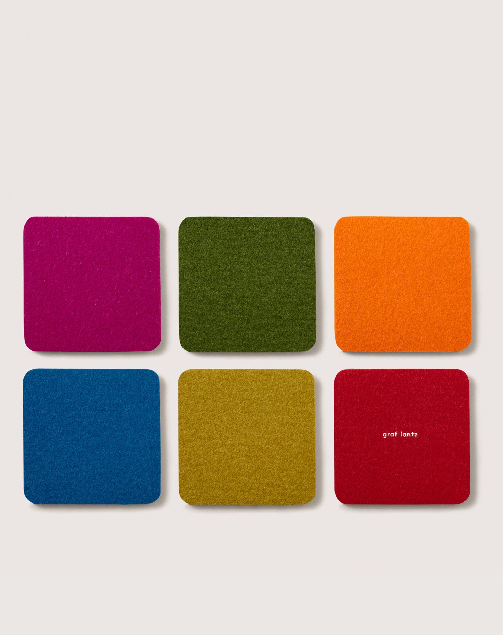 Six square coasters from graf lantz, made of sustainable German Merino wool, come in pink, green, orange, blue, yellow, and mustard. Arranged in two rows on a light background, the second rows right coaster shows graf lantz in white text.